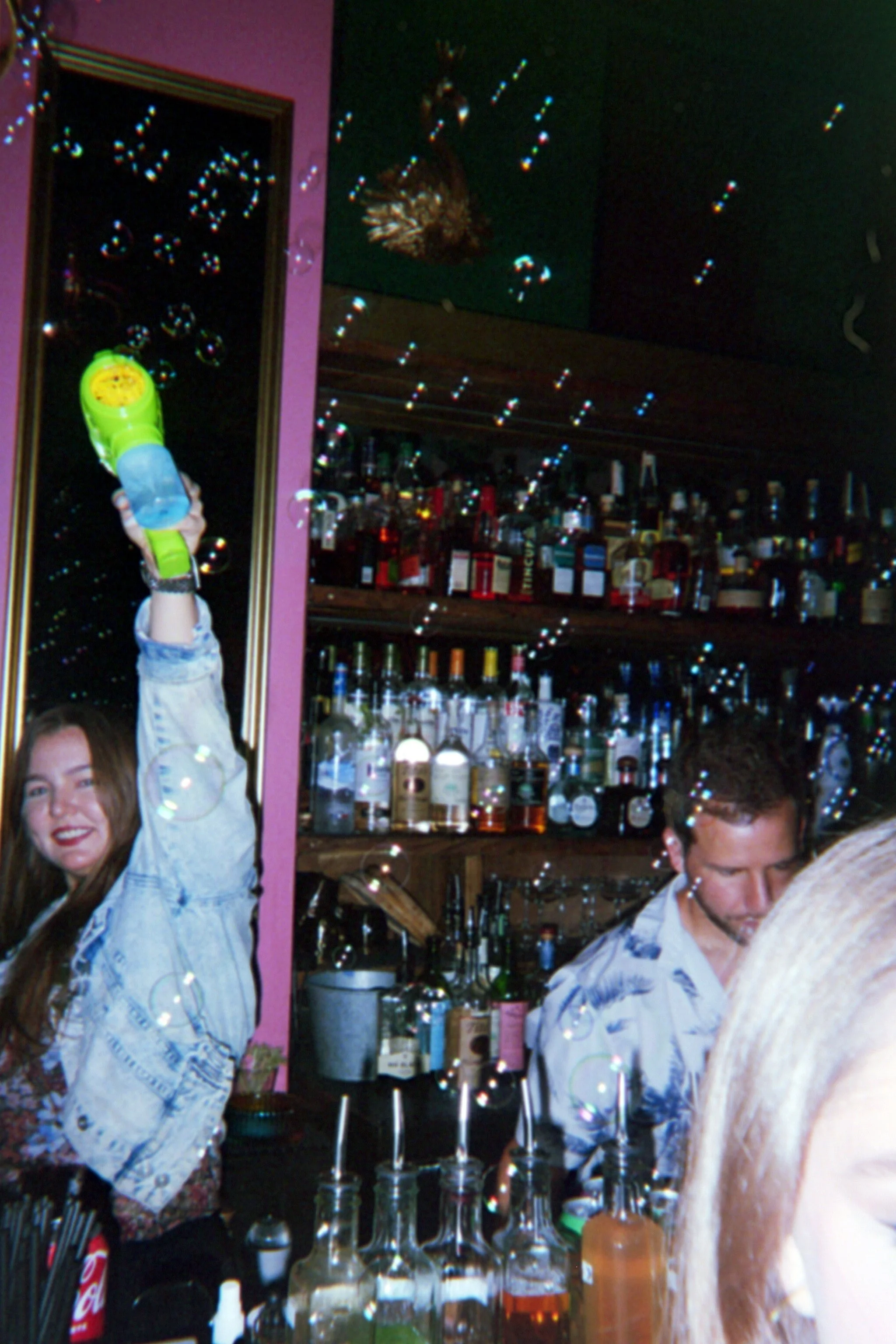 A woman holding a yellow toy water gun in a bar or nightclub with bottles of alcohol on shelves behind her, and bubbles floating in the air.