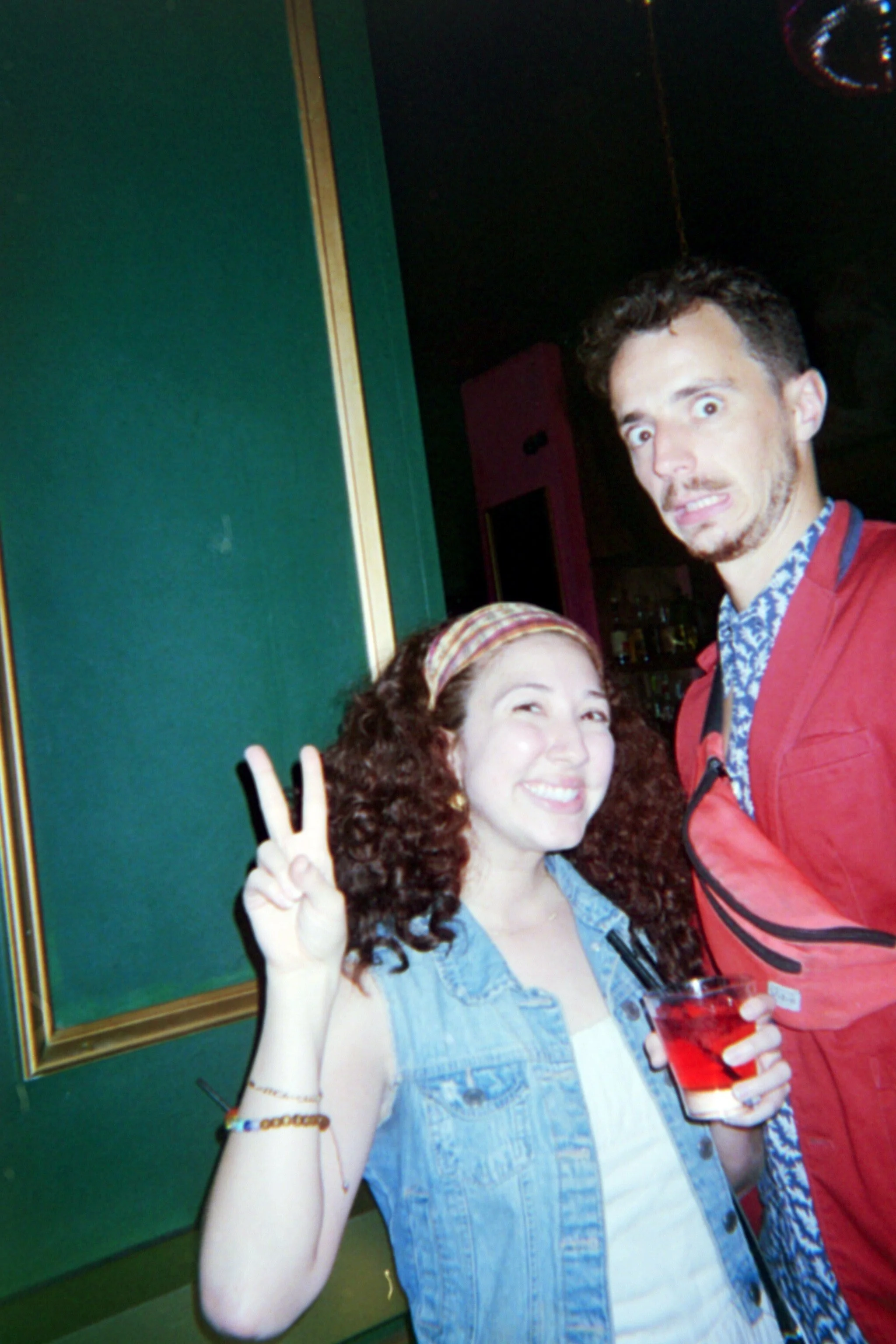 A young woman with curly hair and a headband smiling and making a peace sign, holding a drink. She stands next to a man with a surprised expression in a colorful indoor setting.