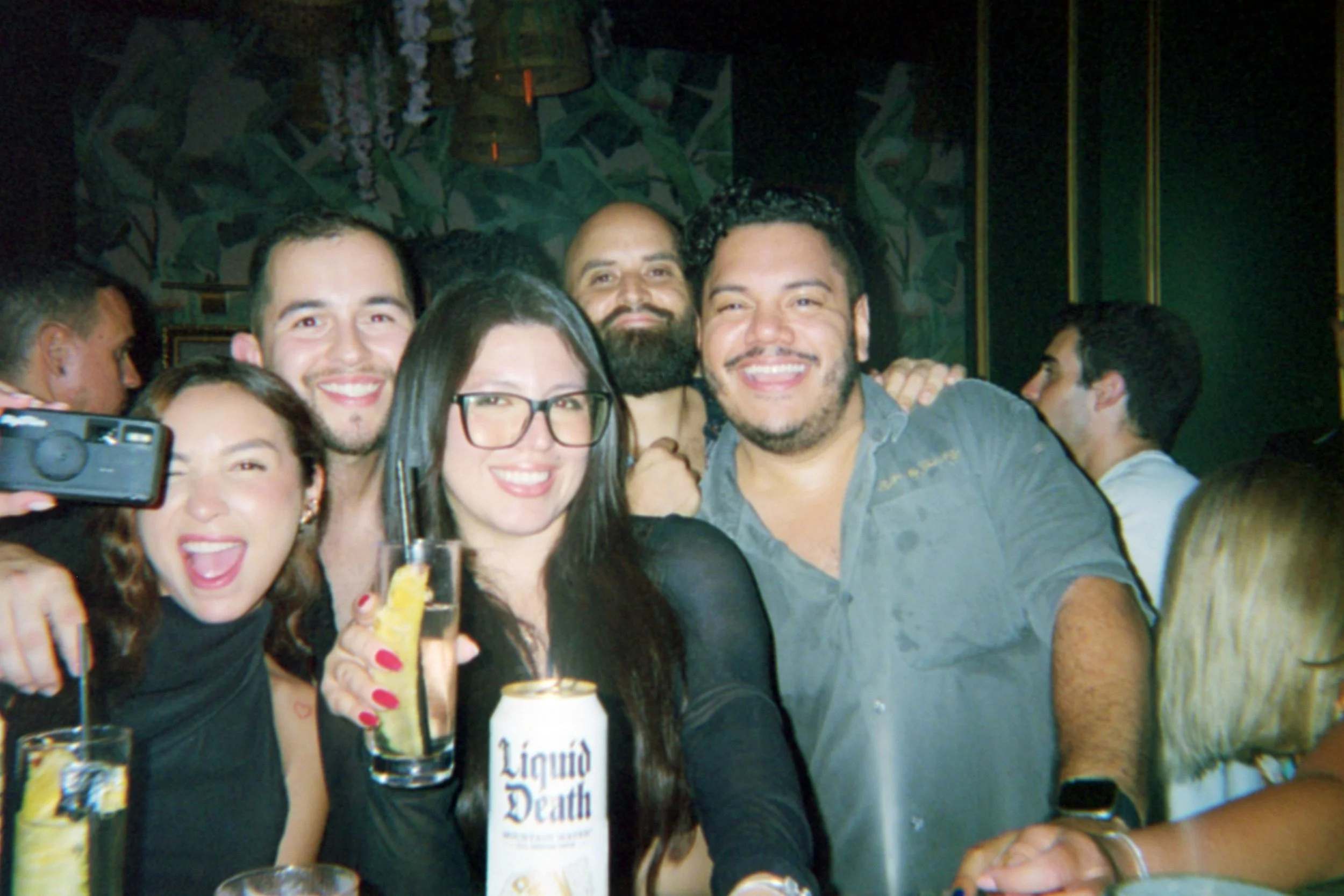 Group of people smiling and celebrating at a party, with drinks in hand, in a dimly lit setting.