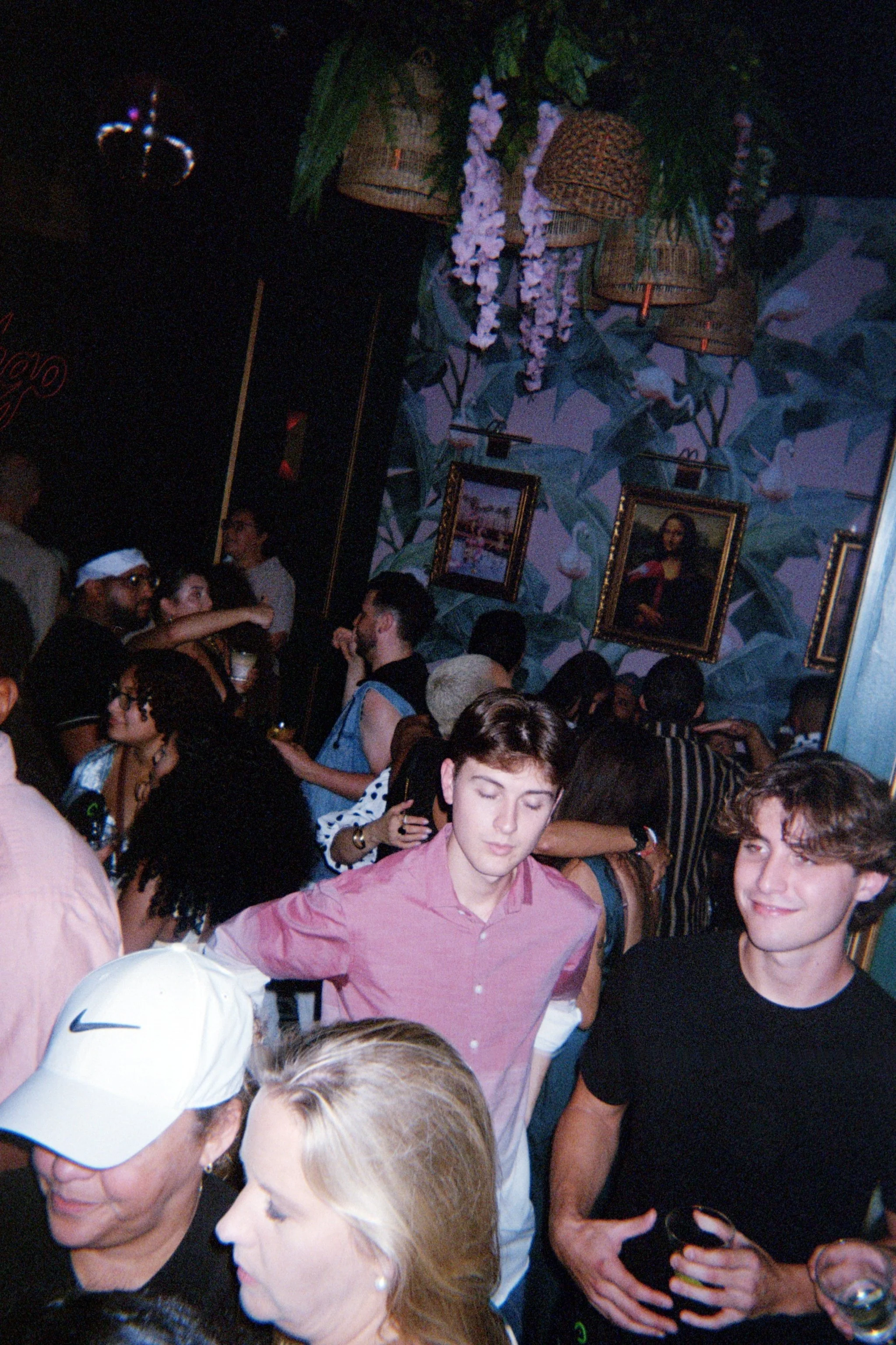 A crowded bar or nightclub with people socializing and drinking, decorated with paintings of the Mona Lisa and tropical foliage on the wall, and woven baskets with hanging flowers.