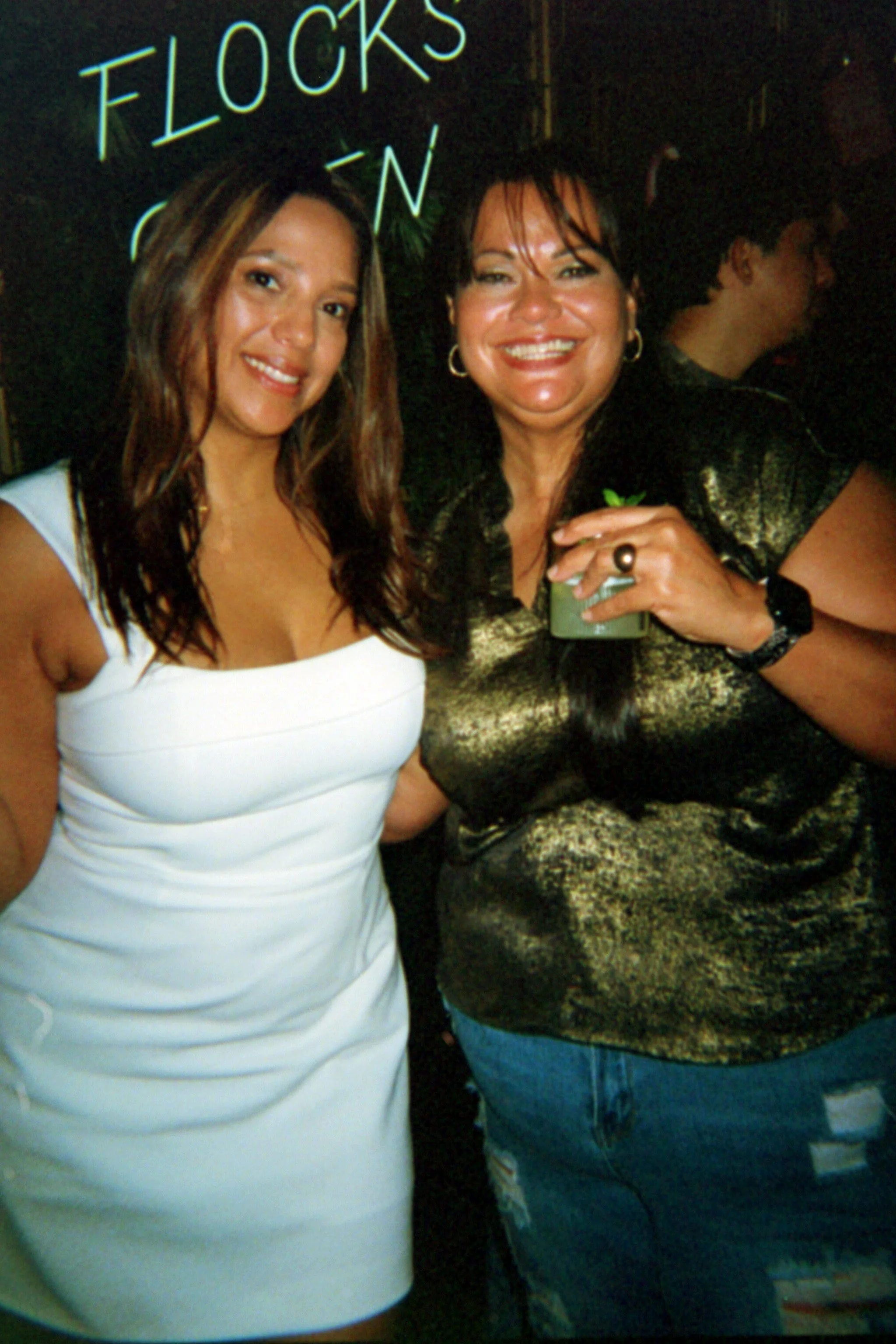 Two women smiling in a dimly lit environment, with a sign reading 'FLOCKS' partially visible behind them.