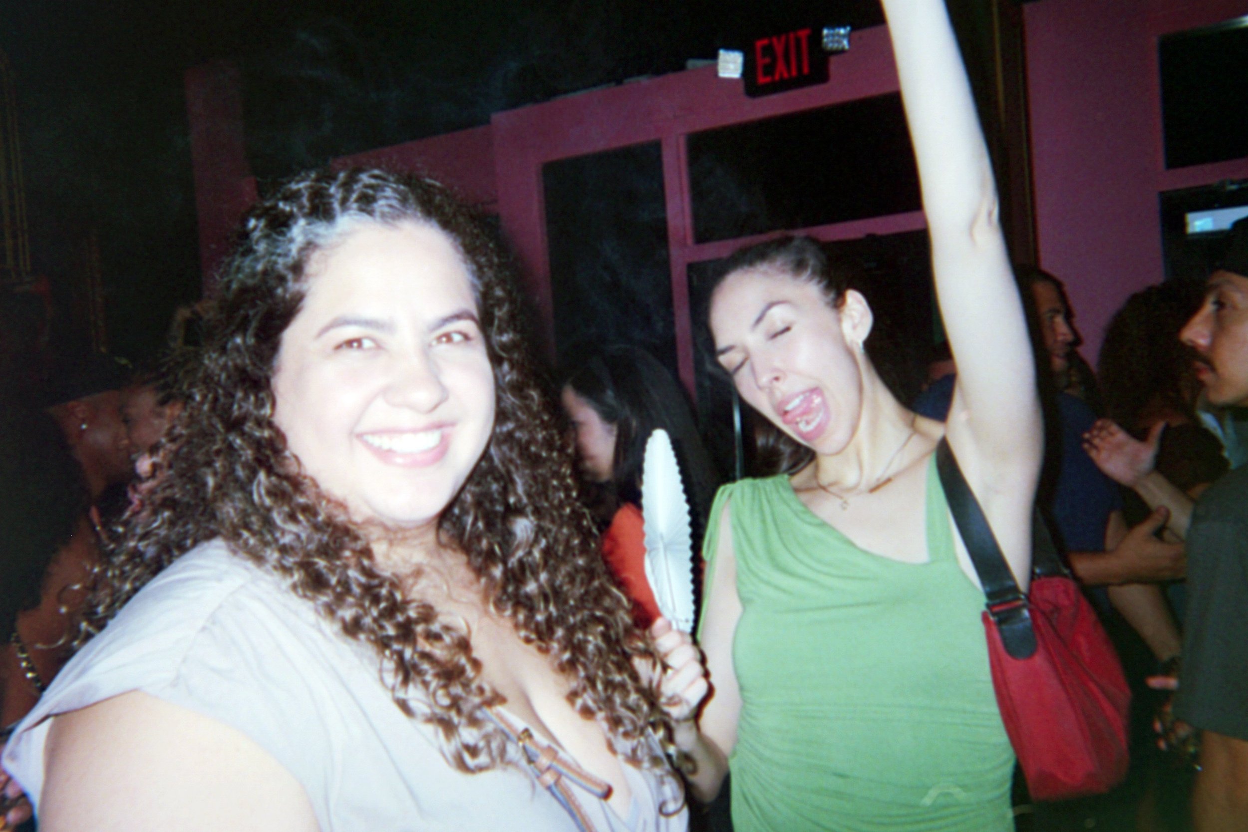 Two women at a social gathering, one smiling at the camera and the other making a playful expression, with people in the background and an exit sign above.