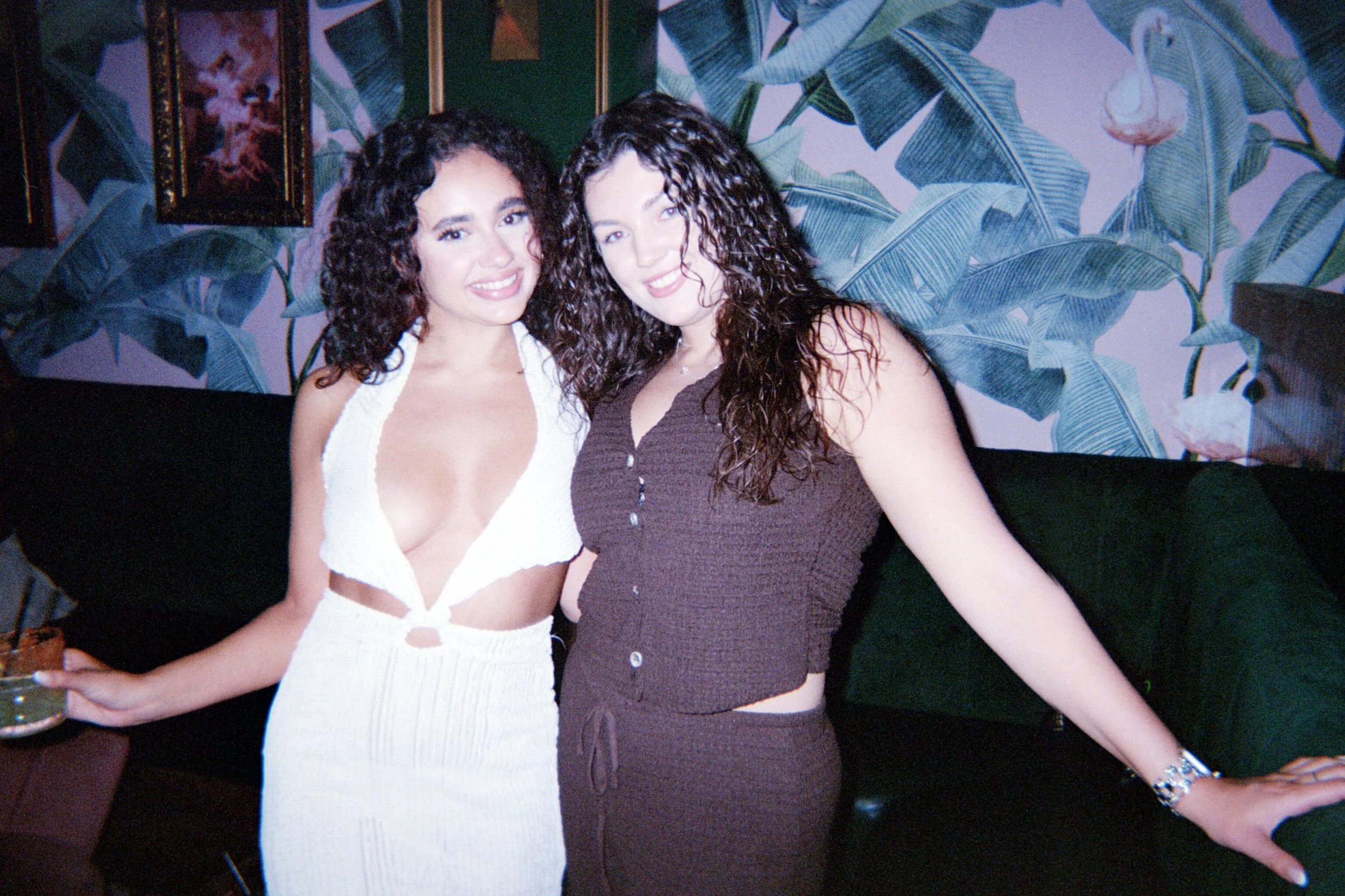 Two women with curly hair smiling and posing together in a room with tropical-themed wallpaper.