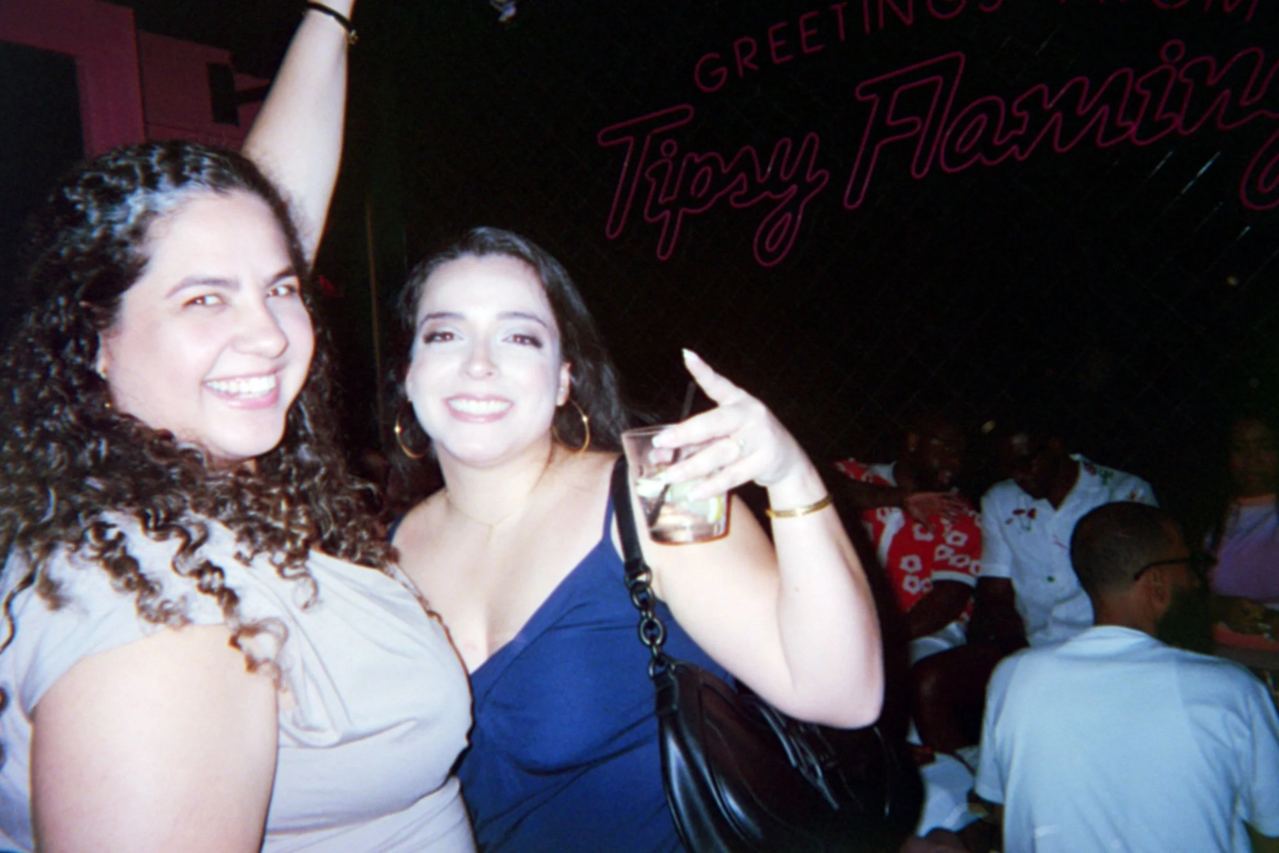 Two women smiling and enjoying a night out, one holding a drink, in a lively venue with a sign that says 'Greetings Tijuana Flamingo' in the background.
