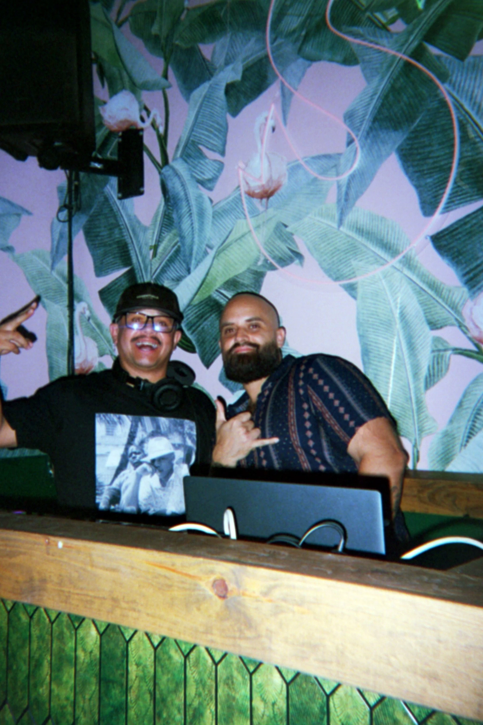 Two men smiling and making hand gestures at a party or DJ booth. One man wears glasses and a dark T-shirt, the other has a beard and a patterned shirt. Behind them is a large wall mural of banana leaves and flamingos.
