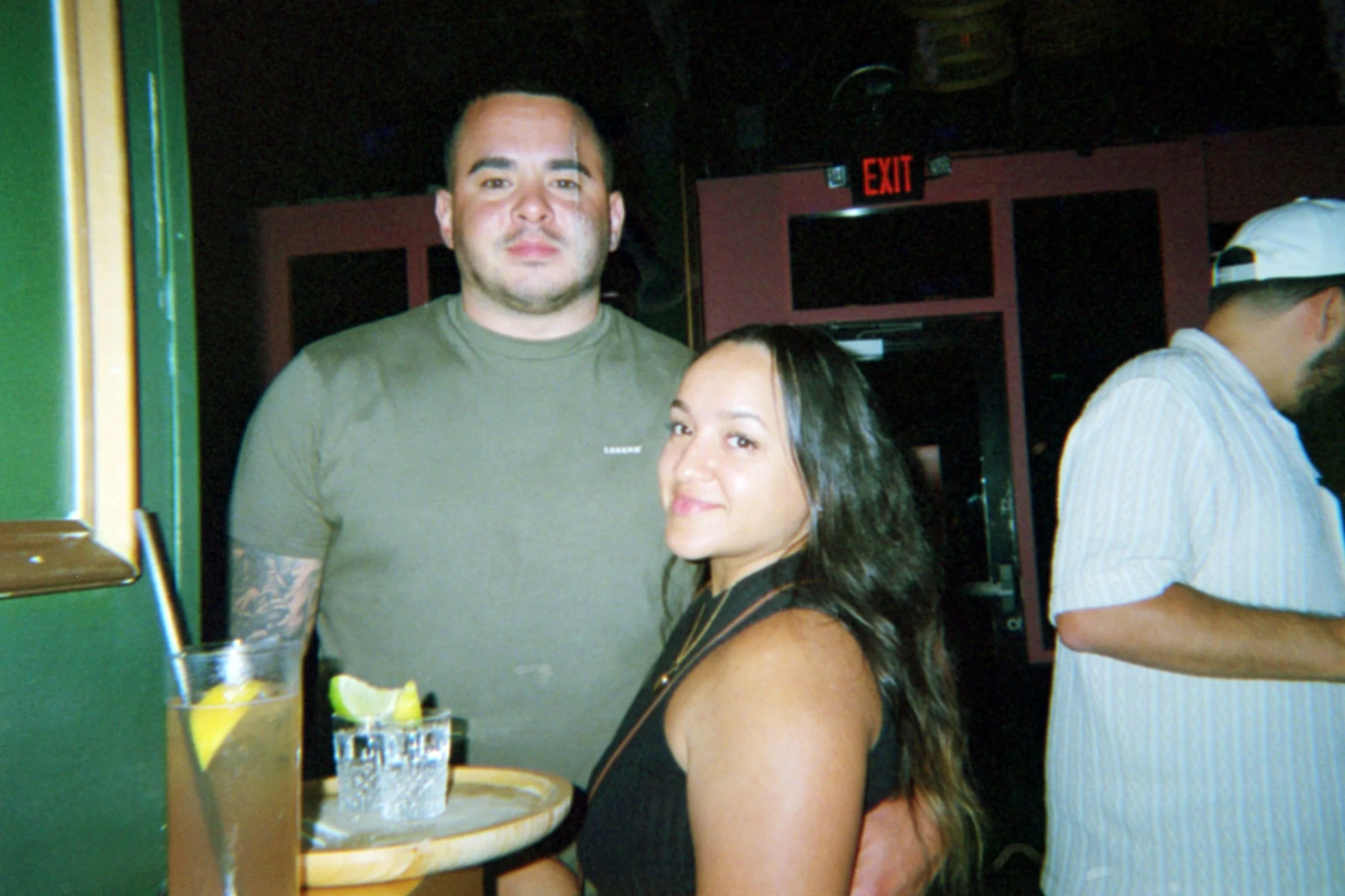 A man and a woman standing close together inside a bar or restaurant, with a drink on a table in front of them, and a man in a white shirt and cap standing nearby.
