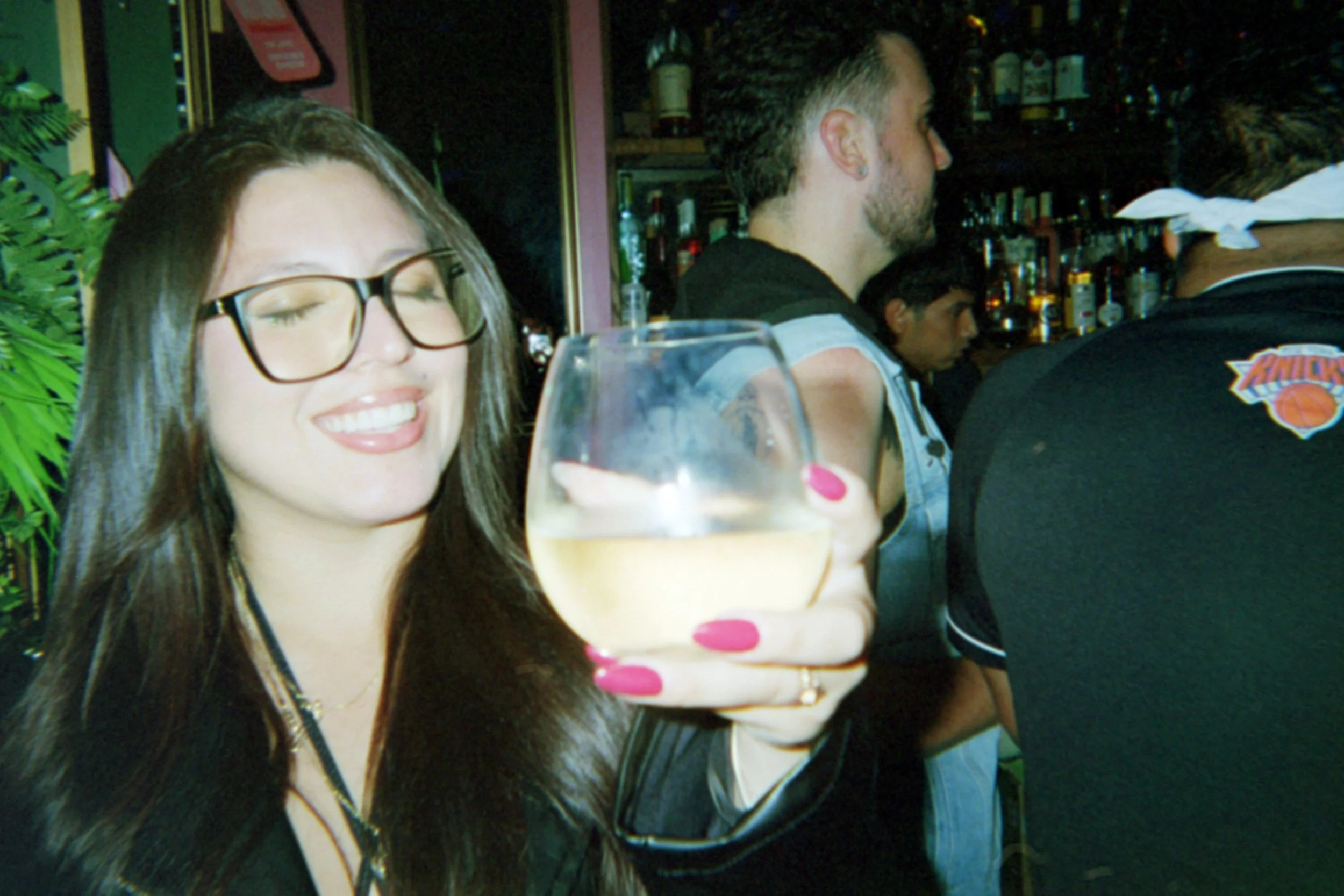 A smiling woman with glasses holding a glass of white wine in a bar or restaurant setting, with other people in the background.