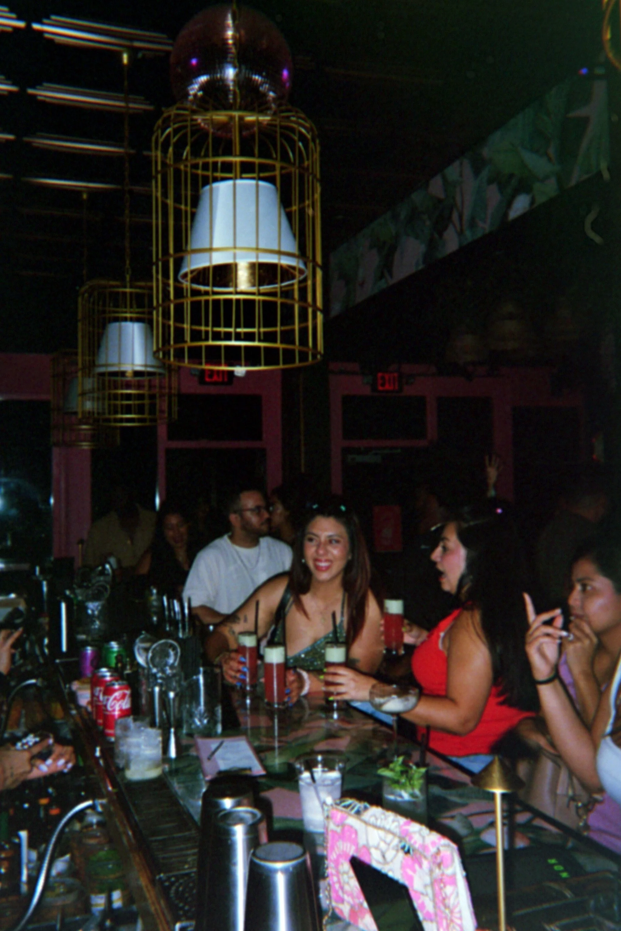 People gathered at a bar, with some holding drinks, in a dimly lit venue featuring hanging lamps with gold accents.