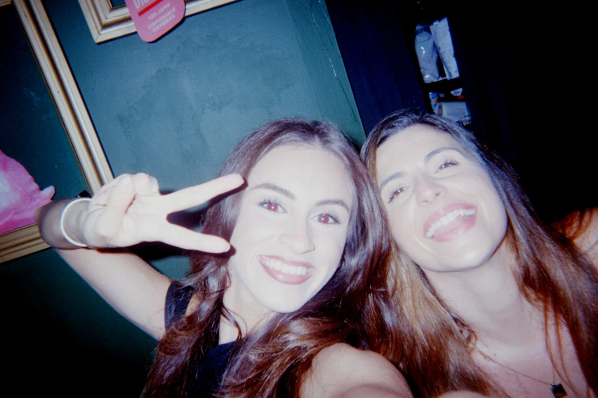 Two women with dark hair smiling and making a peace sign in a dimly lit room.