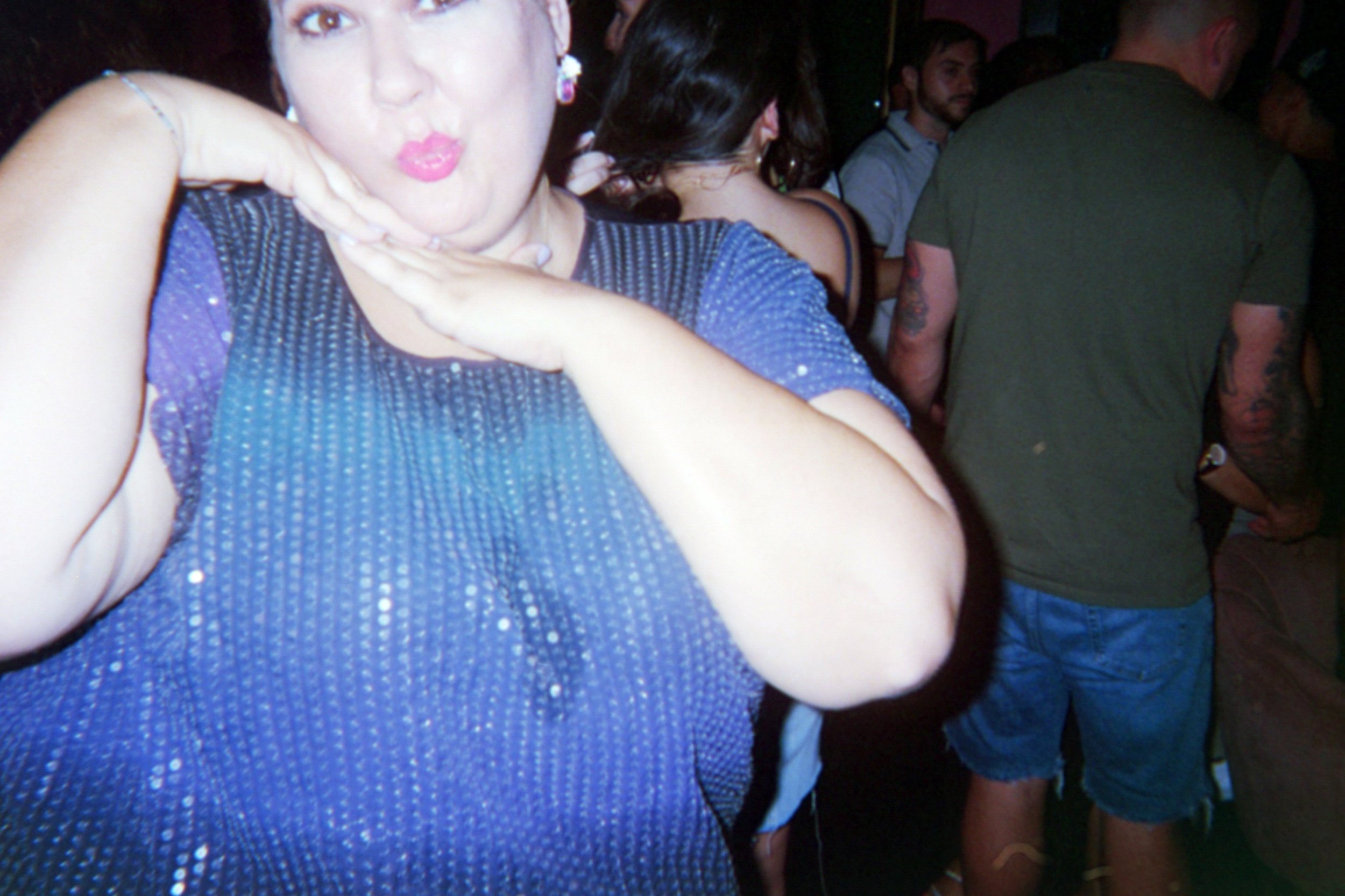 A woman in a blue, sparkly top making a playful facial expression with her hands under her chin in a crowded room.