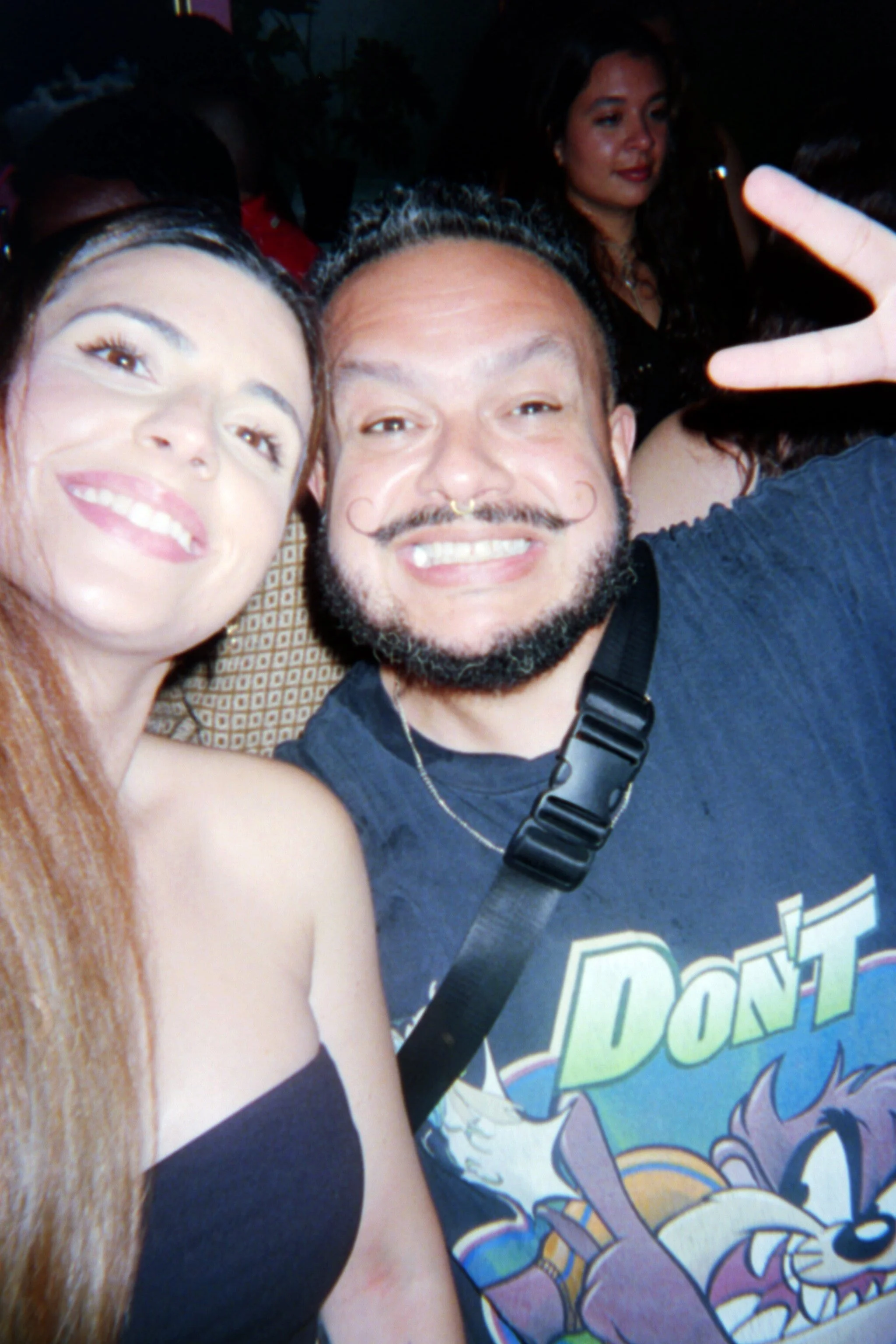 Two people taking a selfie at a social event, smiling with other people in the background.