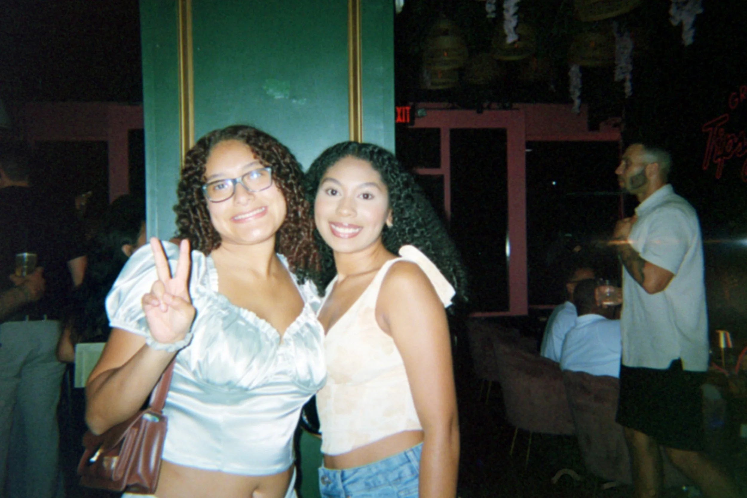 Two women smiling and posing for a photo in a bar or nightclub, one holding up a peace sign, with other people in the background.