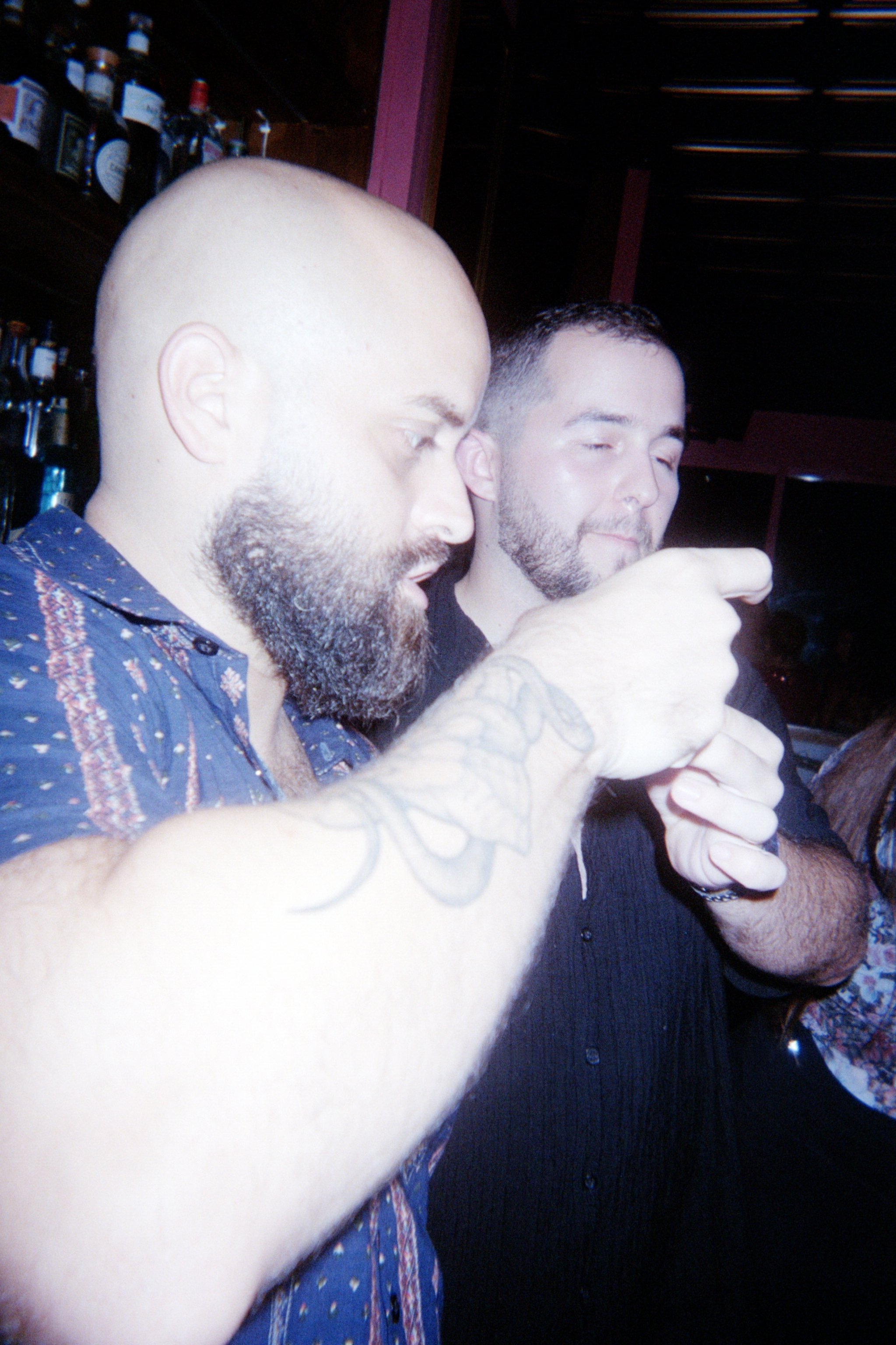 Two men with beards and short dark hair in a bar, one pointing at something while the other looks on, with bottles on shelves in the background.