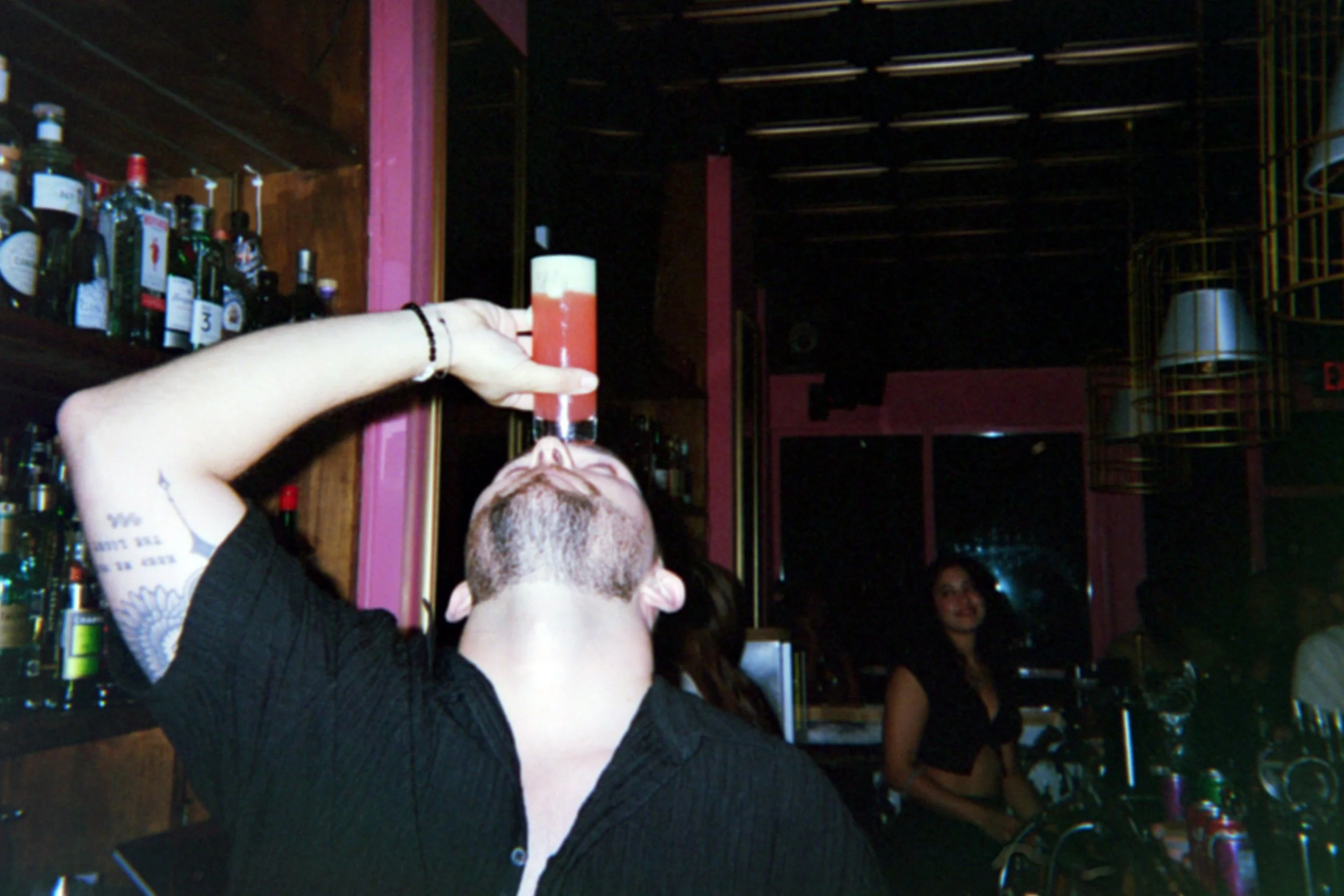 A man in a black shirt with tattoos on his arm is sitting at a bar, tipping a tall glass of red drink with a layer of yellow foam on top, towards his mouth. The bar has shelves with liquor bottles and a woman in a black top is sitting in the backgrou