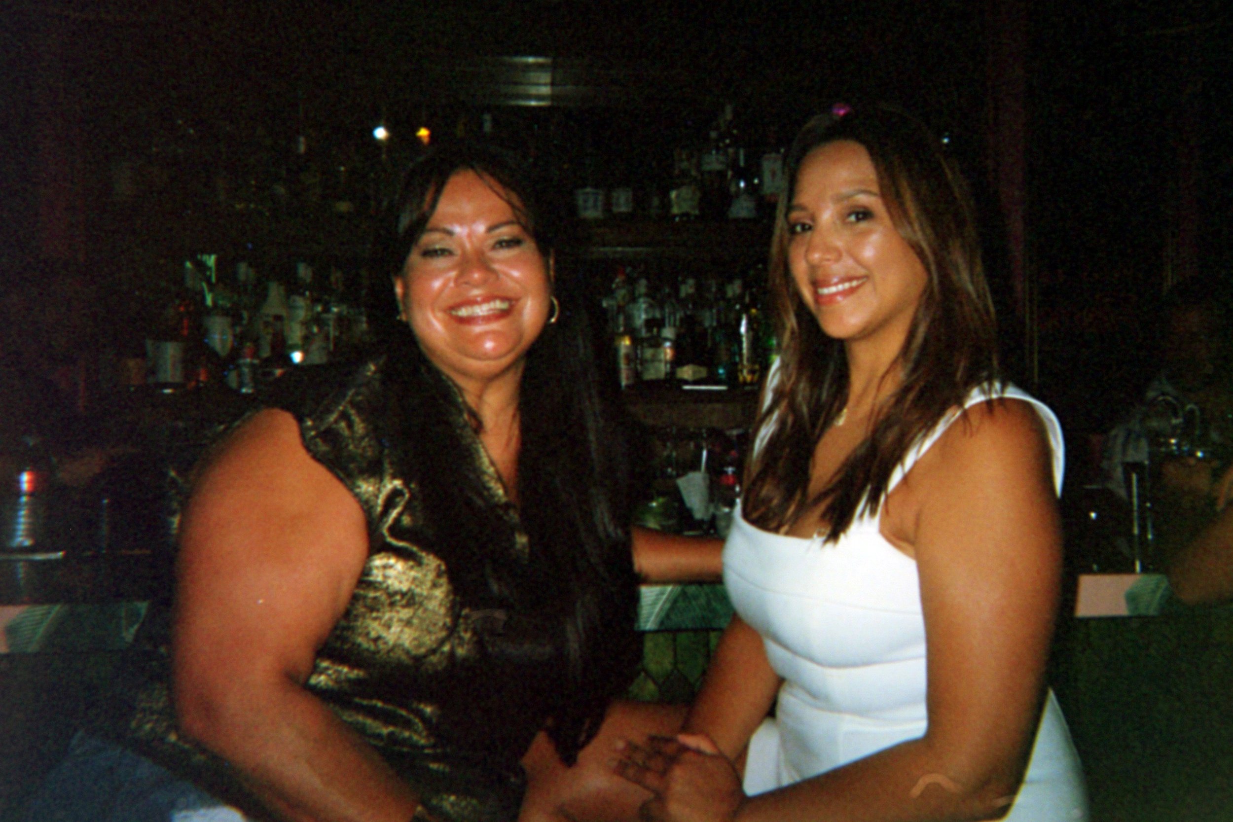 Two women smiling and holding hands in a bar or restaurant setting with shelves of bottles in the background.