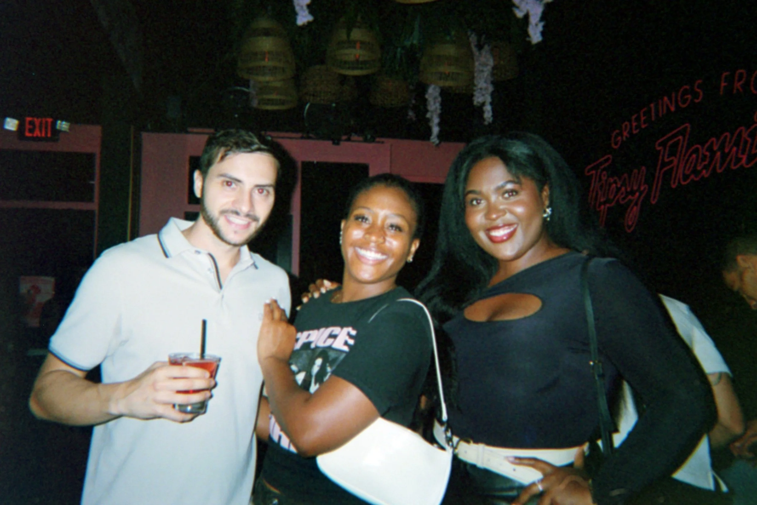 Three people smiling at a bar or club, with two women and one man. The woman on the right has long black hair and is wearing a black long-sleeved top. The woman in the middle has short hair and is wearing a black T-shirt. The man on the left has shor