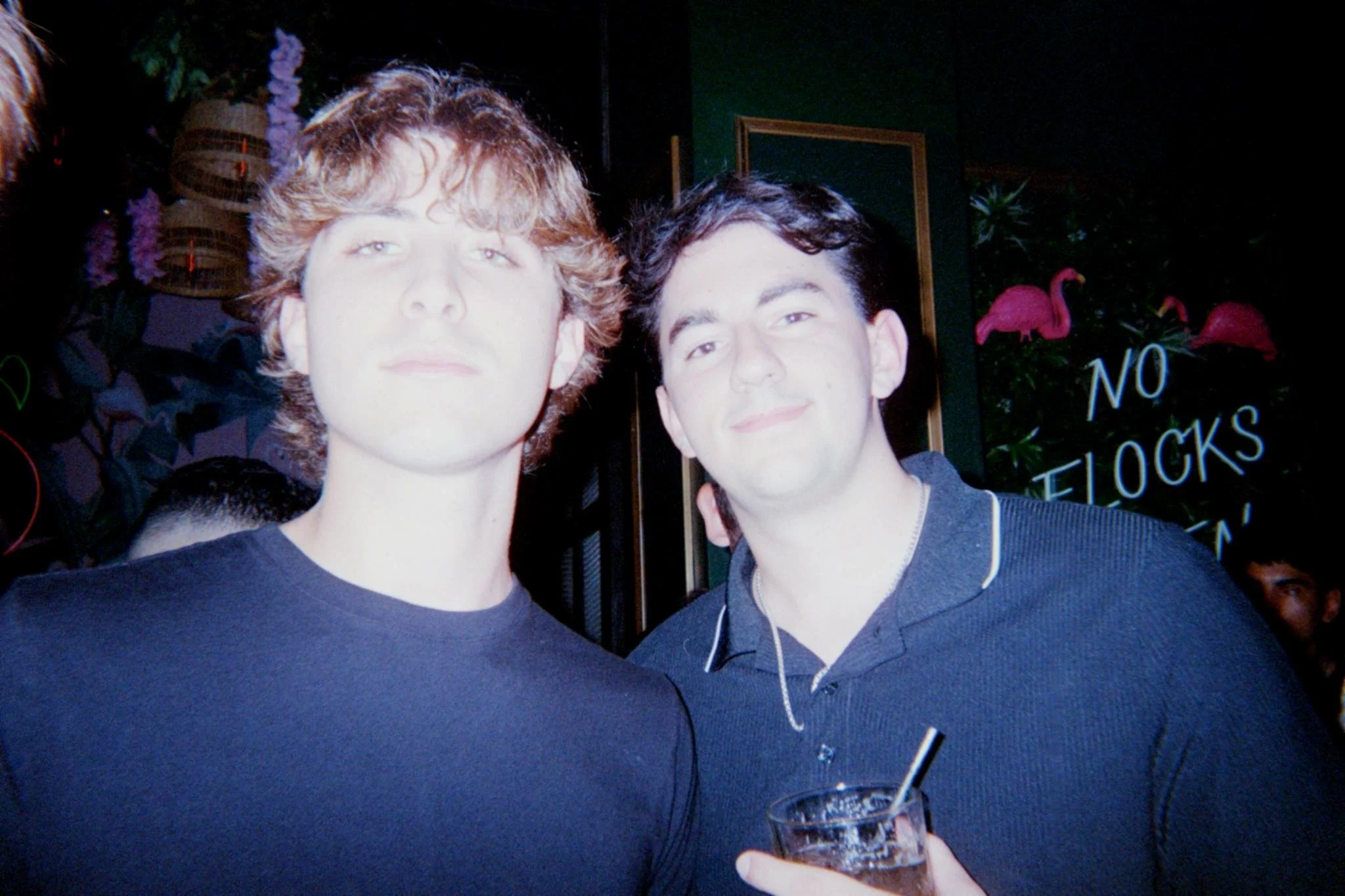 Two young men with dark hair smiling at the camera in a dimly lit venue, one holding a drink with a straw, behind a window with pink flamingo decorations and a sign that reads 'No Flocks'.