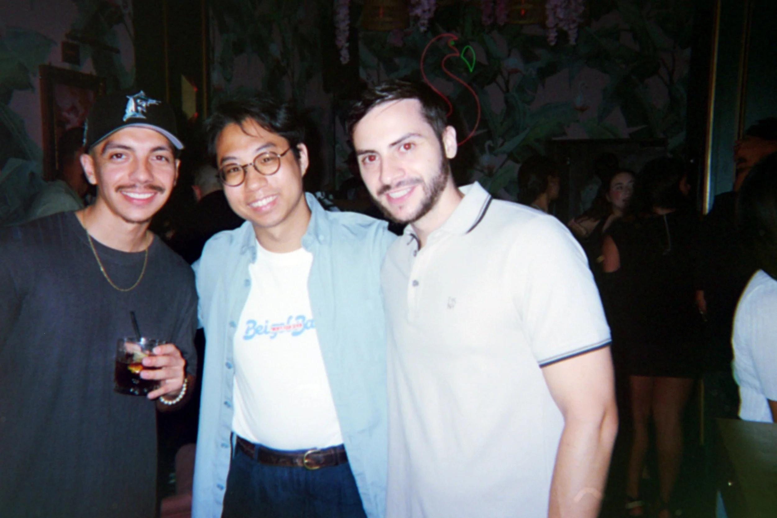 Three men smiling, standing close together at a social event or party, with a dark and colorful background.