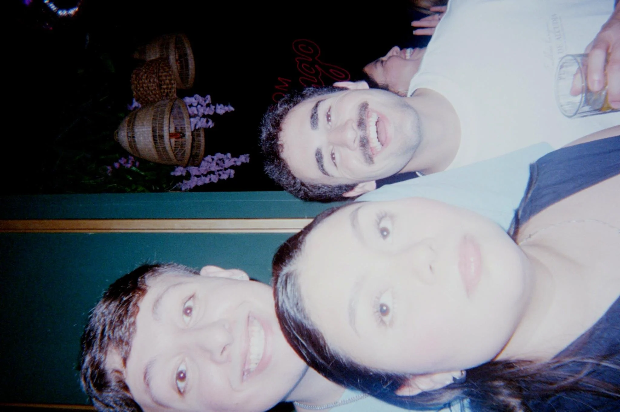 Three people taking a selfie at night, with a dark background featuring some decorative baskets and flowers.
