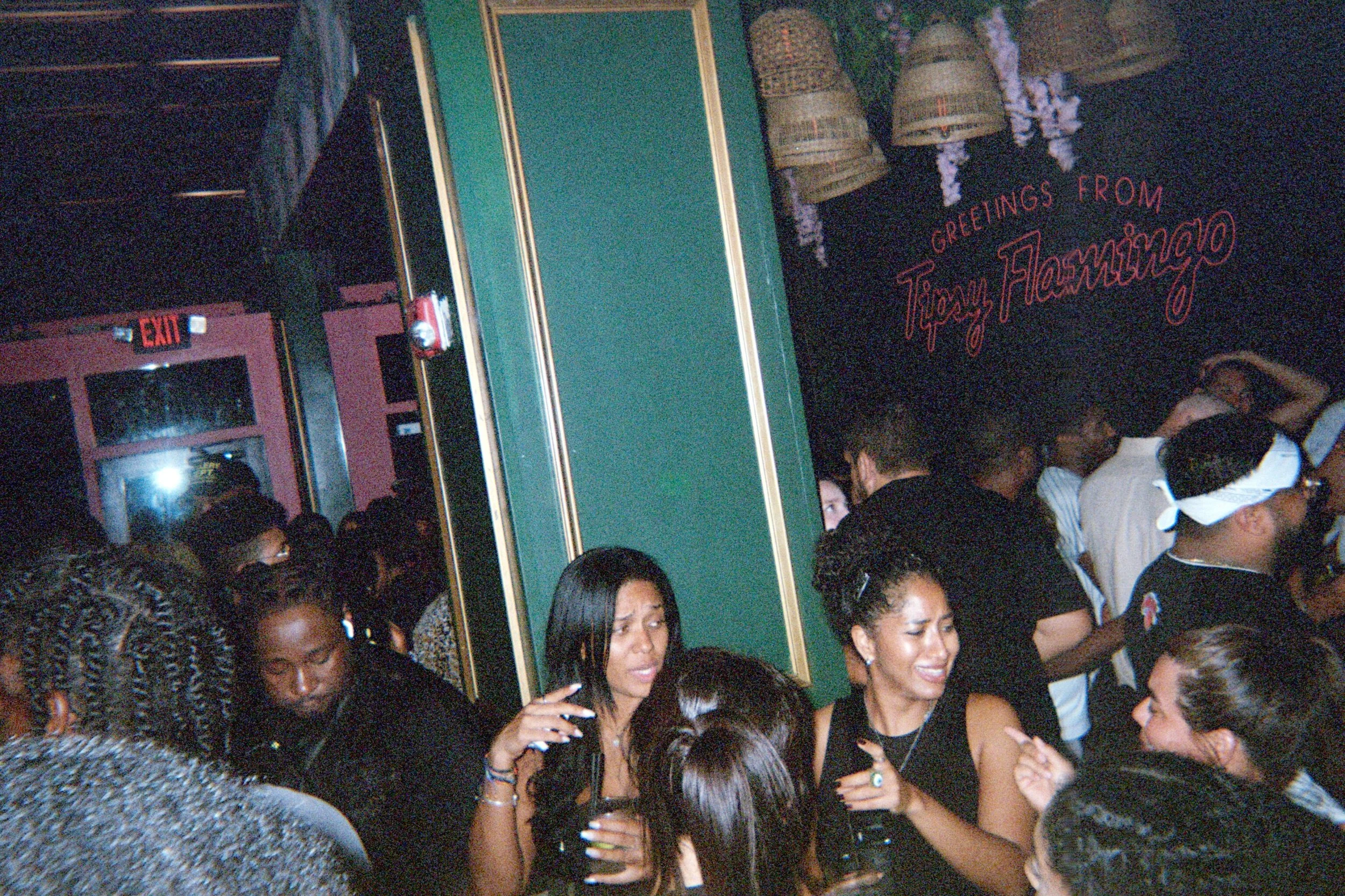 Crowd of people socializing in a dimly lit bar or club, with a neon sign on the wall that reads 'Greetings from Tippy Flamingo' and decorative hanging baskets.