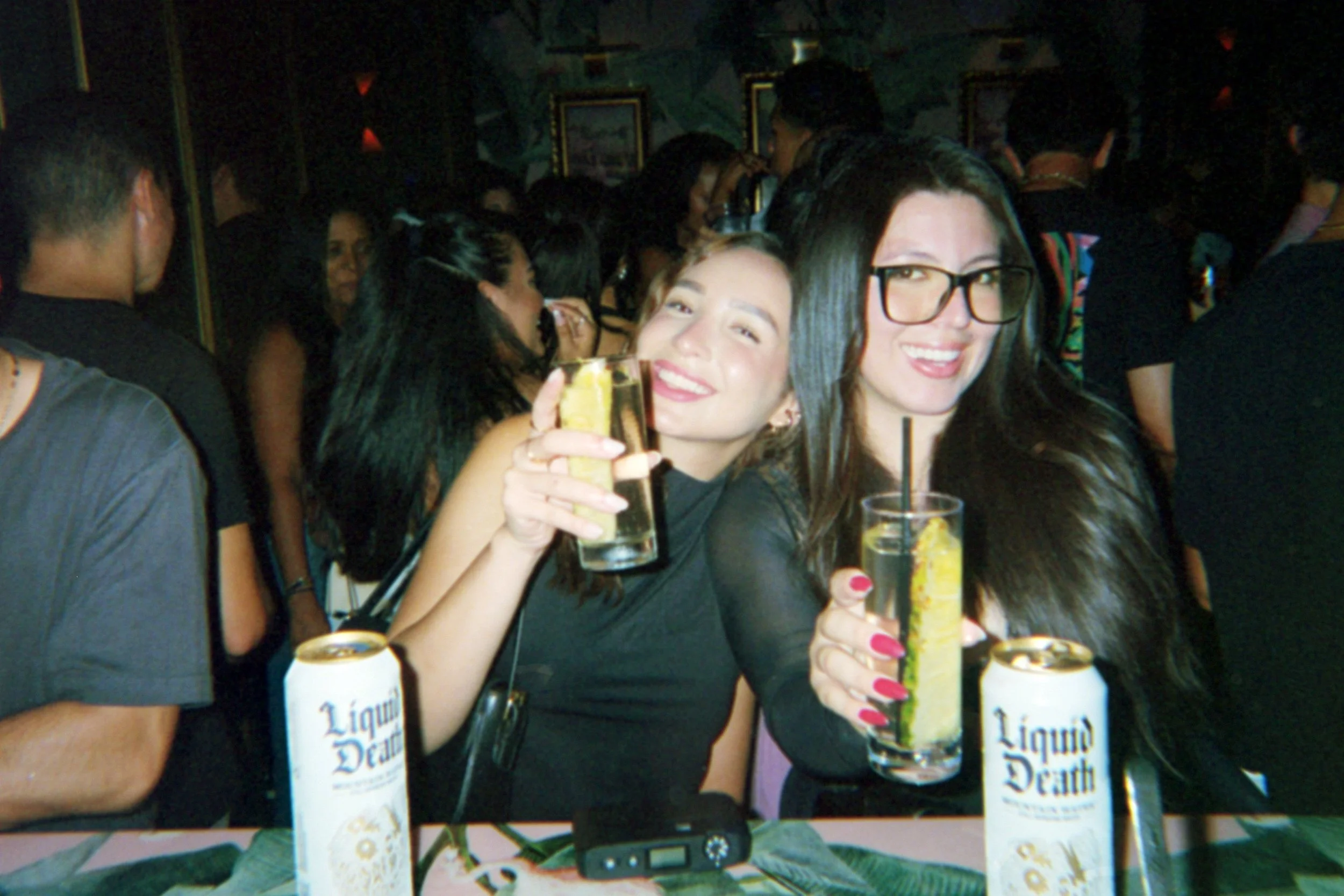 Two women smiling and holding drinks at a crowded bar or party, with cans of Liquid Death water and a smartphone on the table.