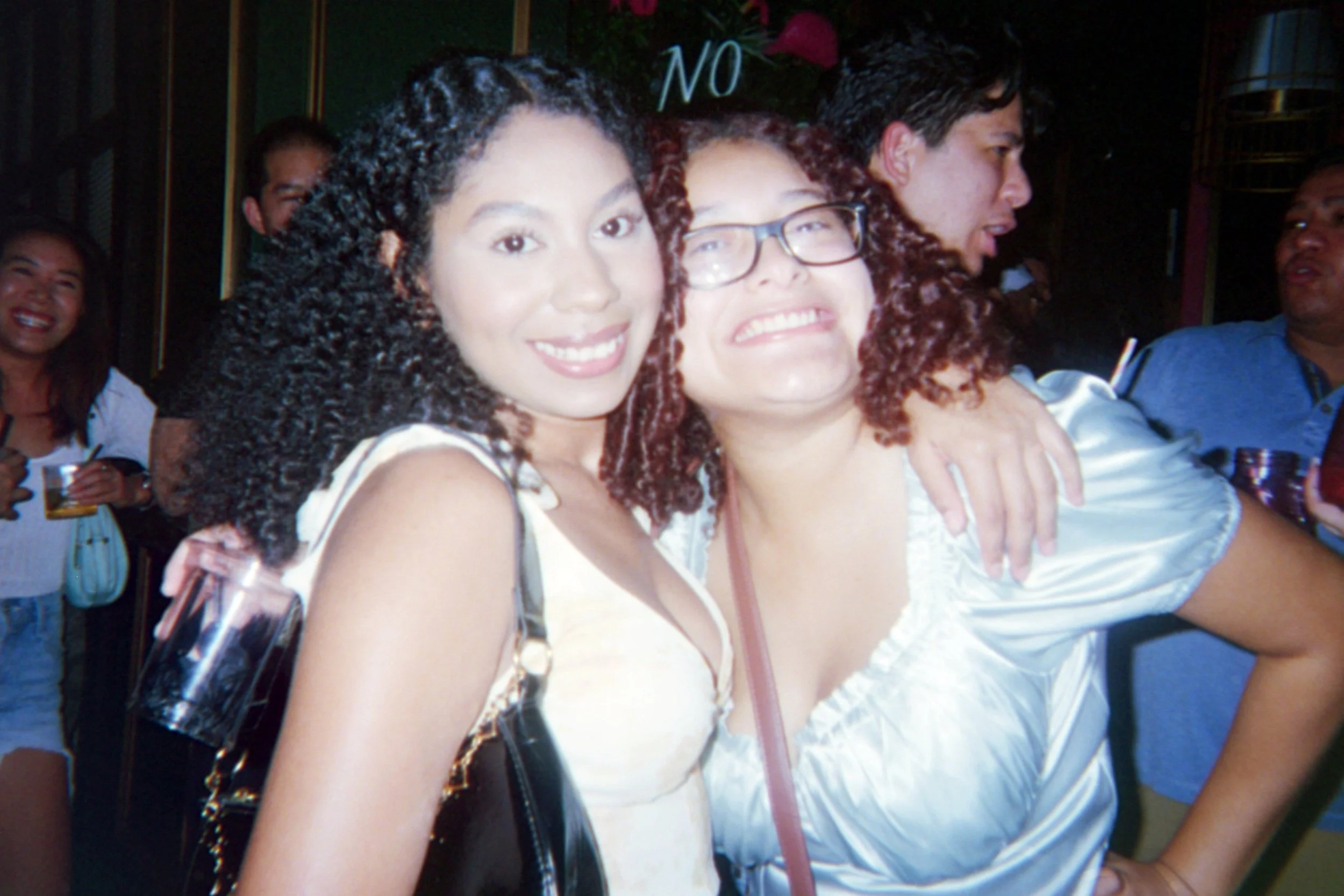 Group of smiling women and men at a social gathering, two women in the foreground are embracing and smiling at the camera.