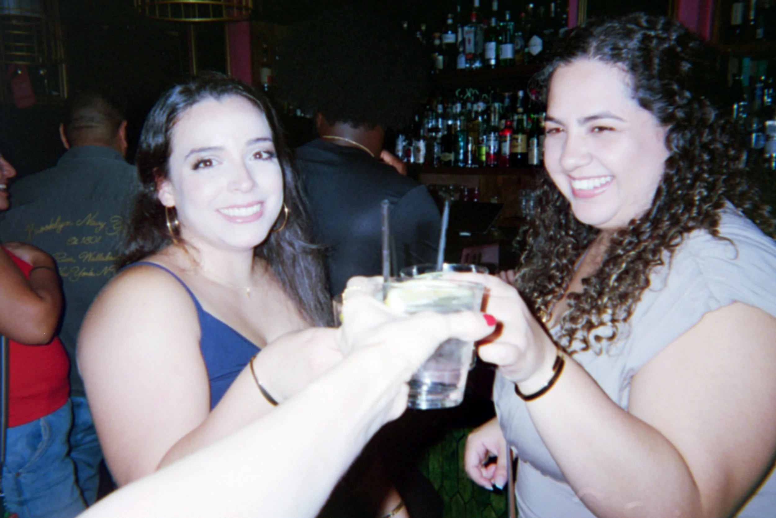 Two women in a bar raising glasses for a toast, smiling, with a background of a bar stocked with bottles.