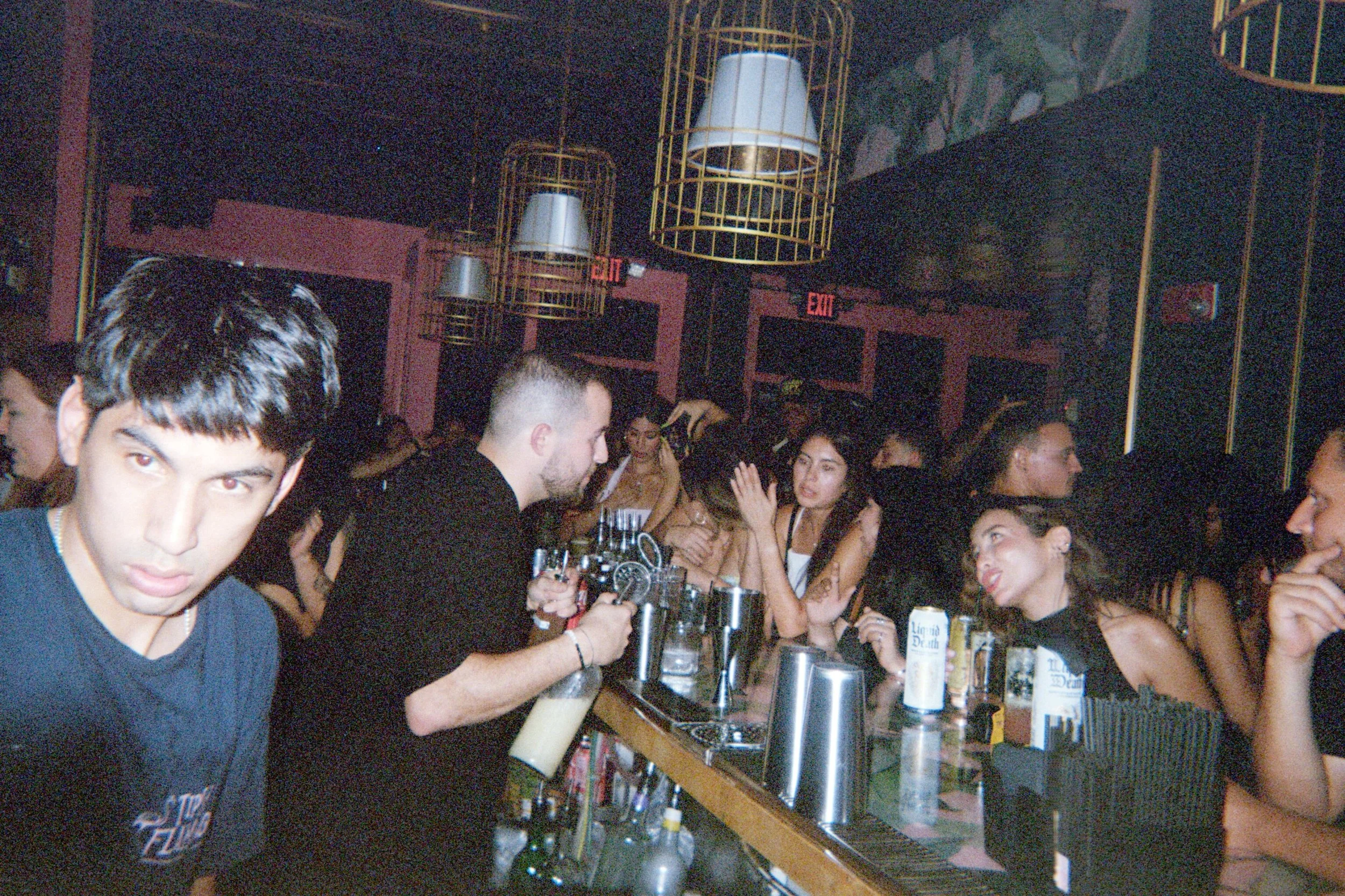 People socializing at a bar with drinks, in a dimly lit nightclub or bar setting.