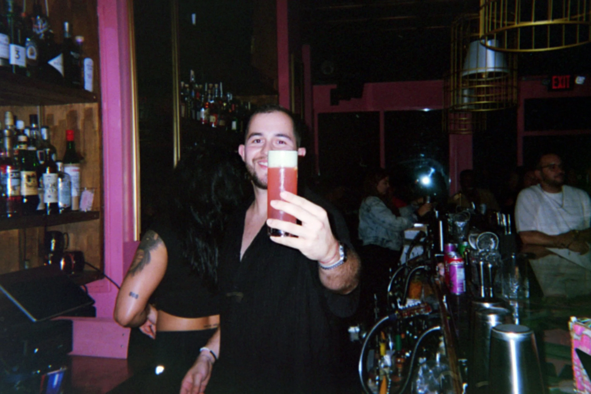 A man with dark hair and beard holding a tall glass of beer in a bar. The bar has shelves with bottles and other patrons in the background.