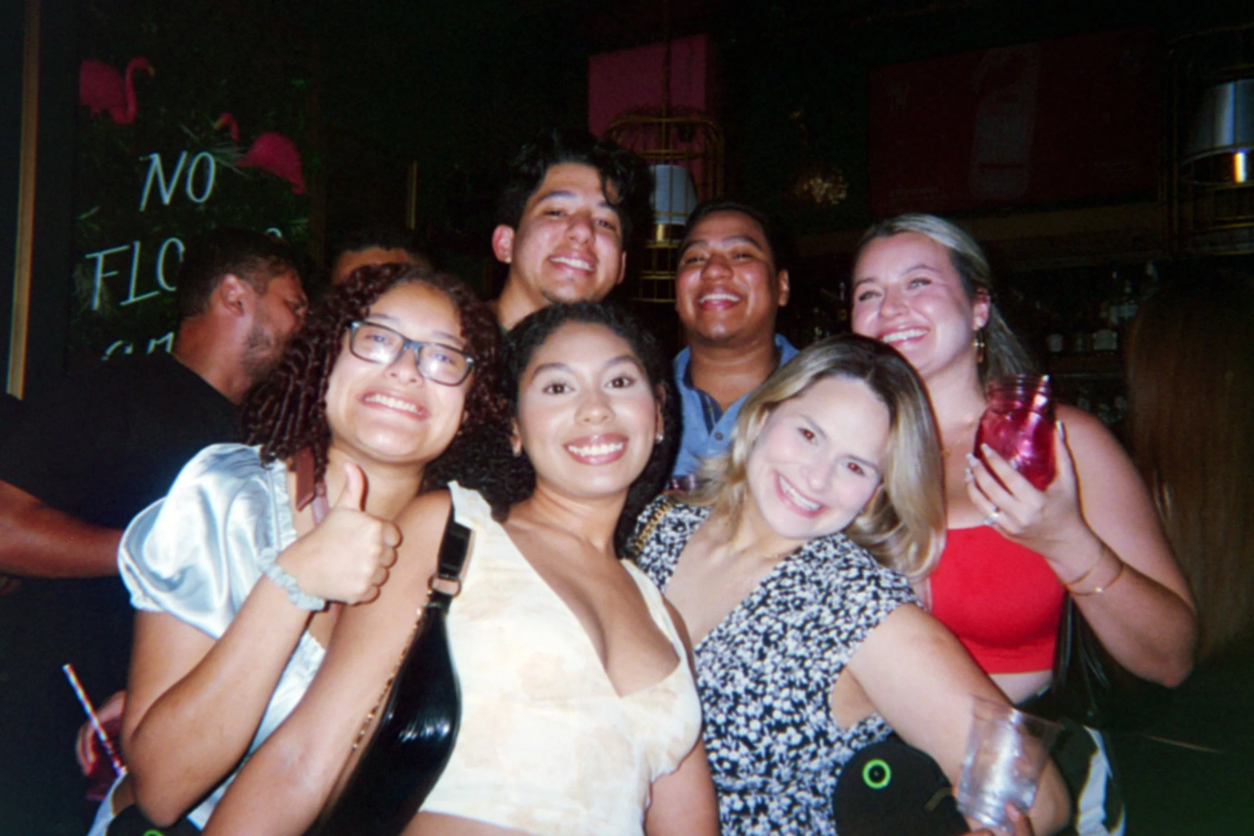 A group of seven diverse friends smiling and posing together at a social gathering in a dimly lit venue, with one person giving a thumbs-up and another holding a drink.