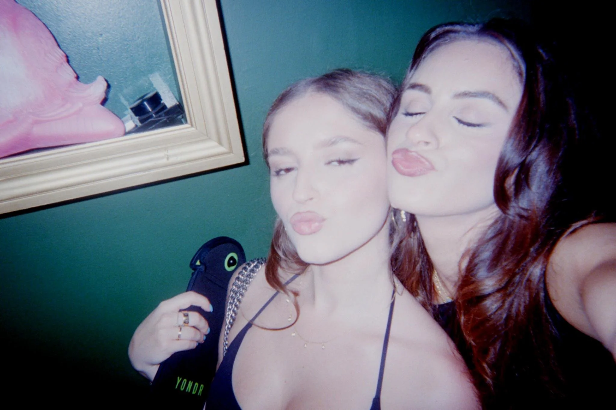 Two women taking a selfie. One has light brown hair and the other has dark brown or black hair. Both are making kissy faces. The background includes a mirror and a green wall.