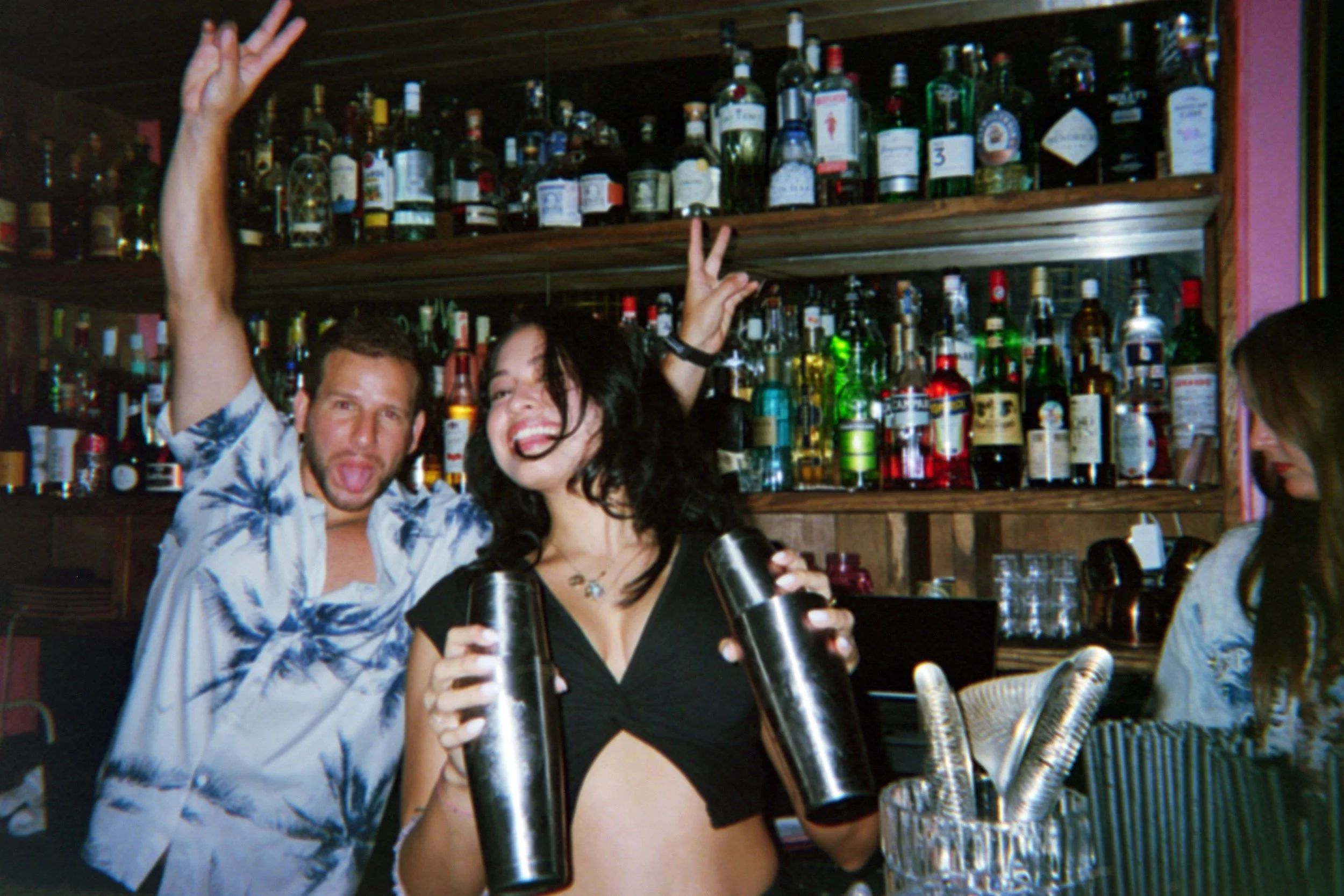 Two people celebrating at a bar, with one man and one woman holding cocktail shaker bottles. They are smiling and making peace signs, with a large selection of liquor bottles on shelves behind them.