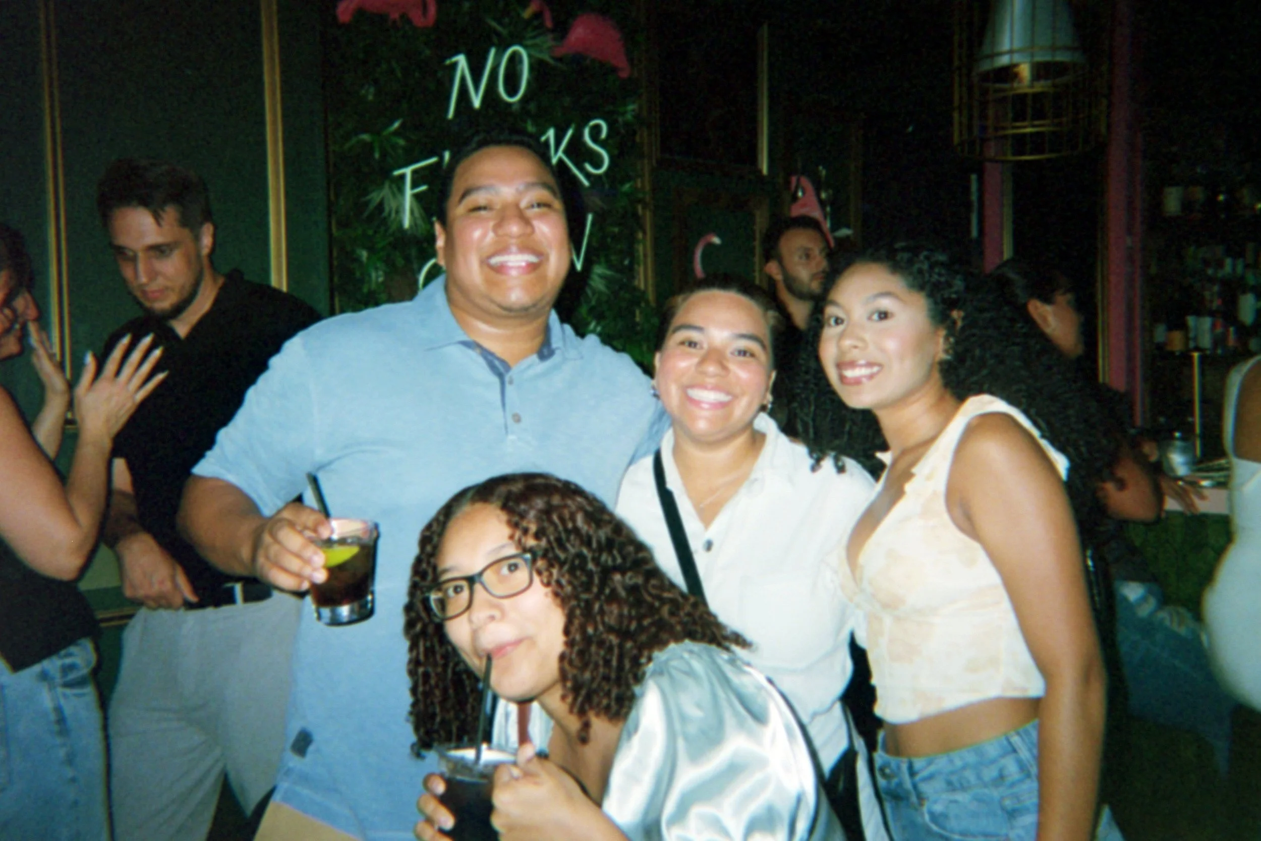 Group of five friends at a bar or nightclub, smiling and posing for a photo. Two women are holding drinks, one man is holding a drink, and they are standing close together. The background has neon signs and decorations.