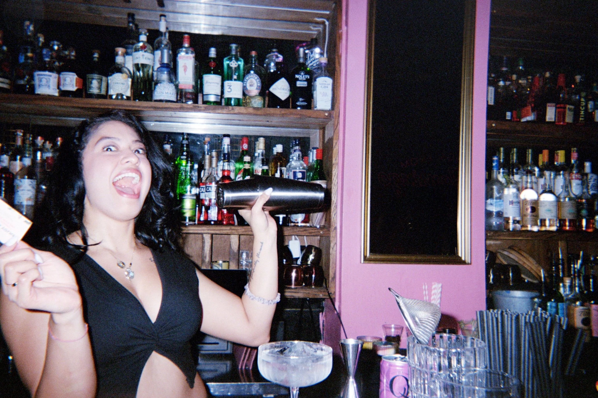 A woman with dark hair, wearing a black top, behind a bar counter, smiling, holding a cocktail shaker, with bottles of alcohol on shelves in the background.