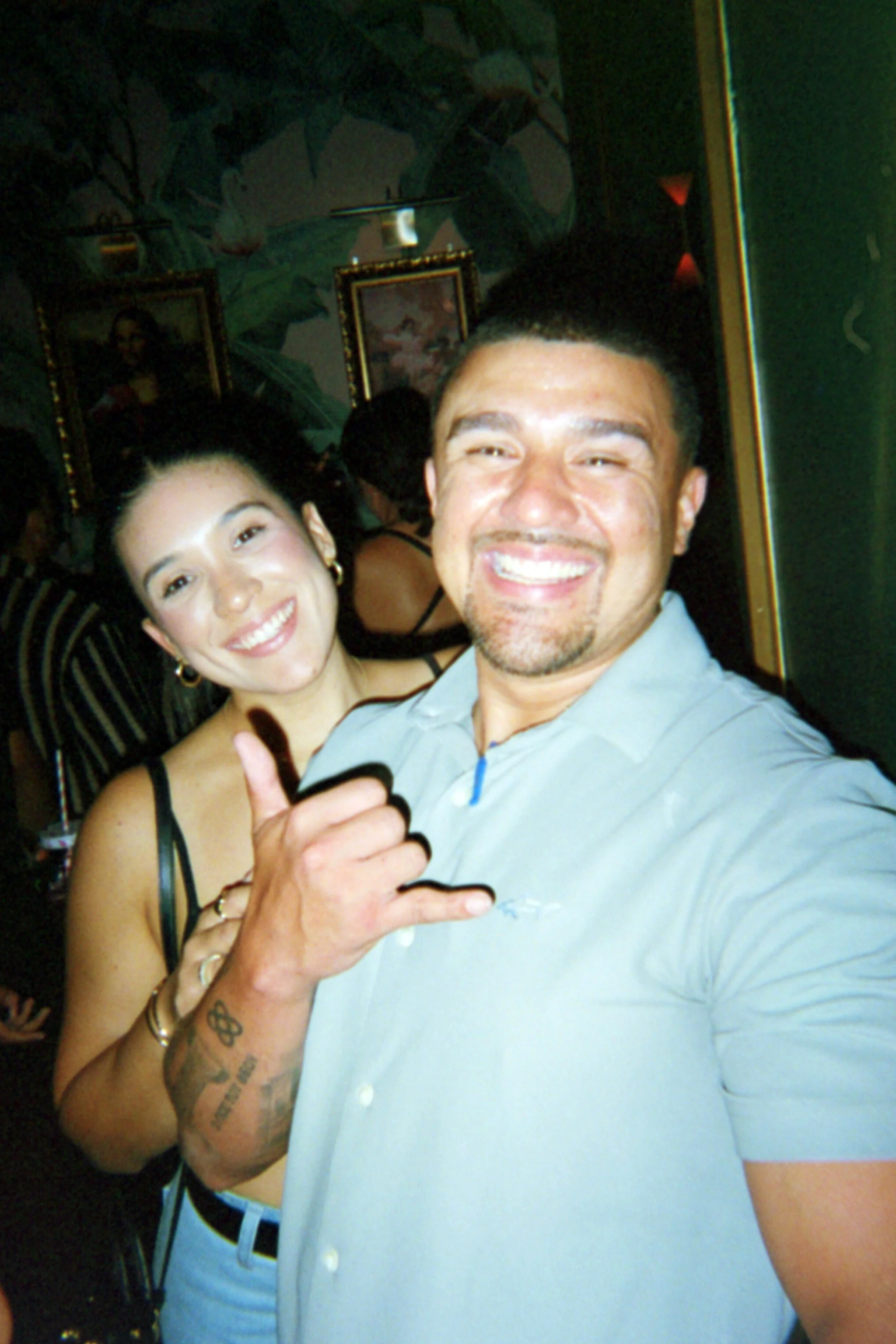 Two smiling friends taking a selfie at a social gathering, with one making a shaka hand sign.