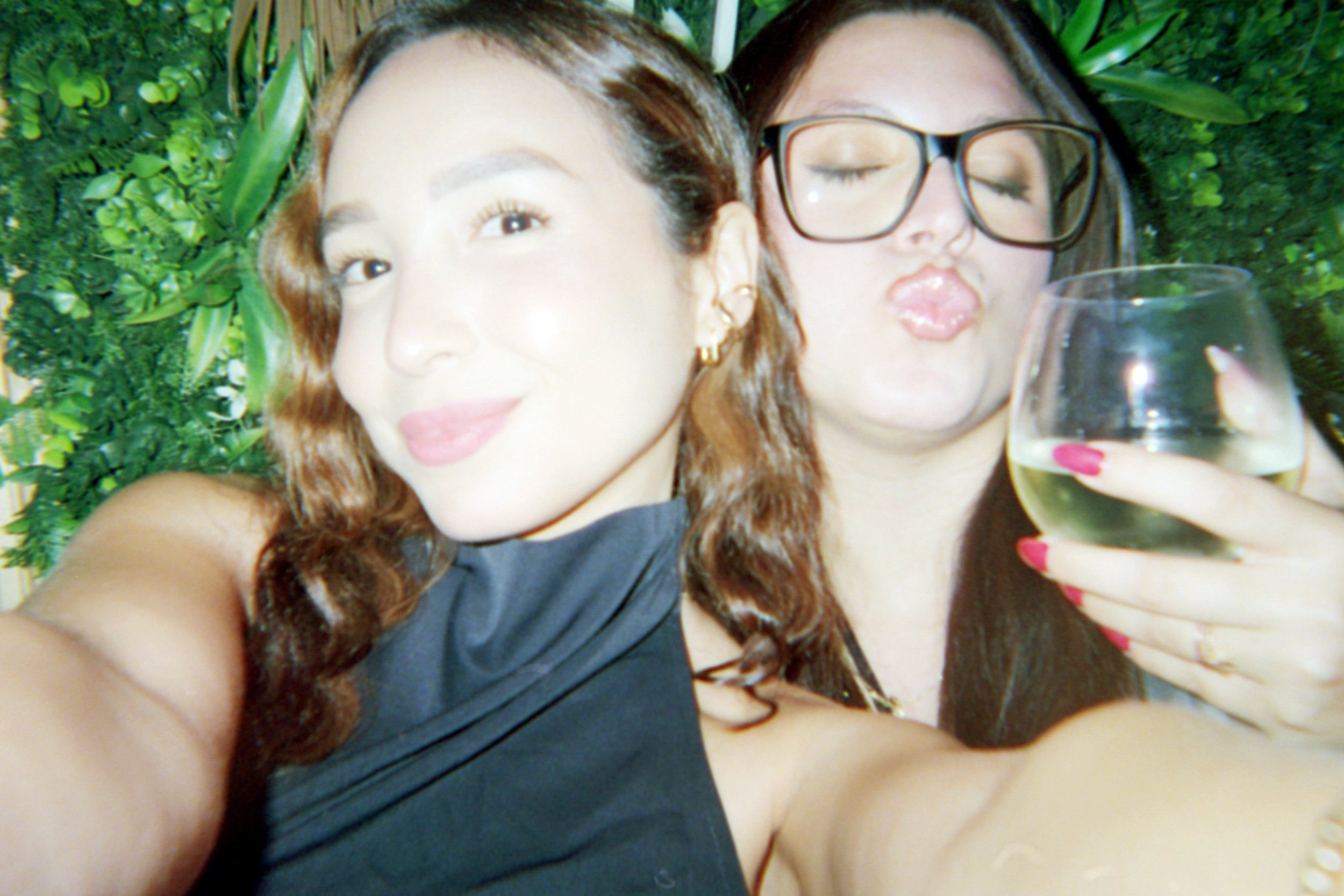 Two women taking a selfie together, one with light skin and wavy hair, smiling, and the other with darker skin, glasses, and pursing lips, holding a glass of white wine, against a green leafy background.