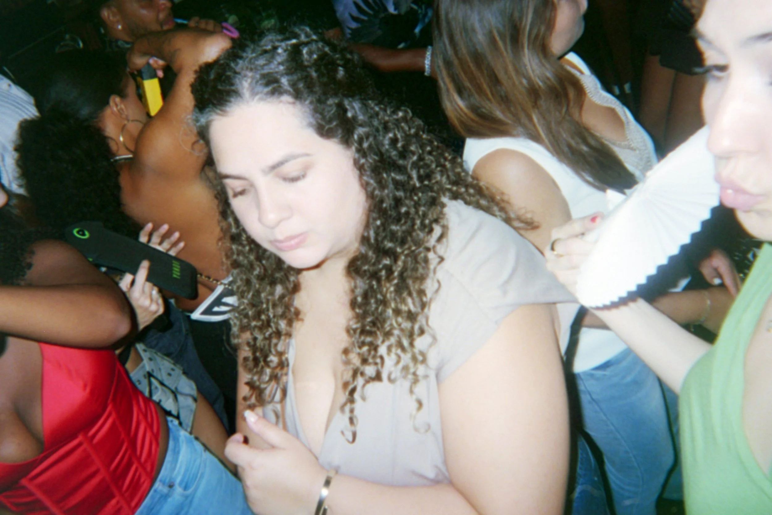 Crowded scene of diverse women standing closely together, some using their phones and holding umbrellas, with a prominent woman with curly hair and a deep neckline in the center.