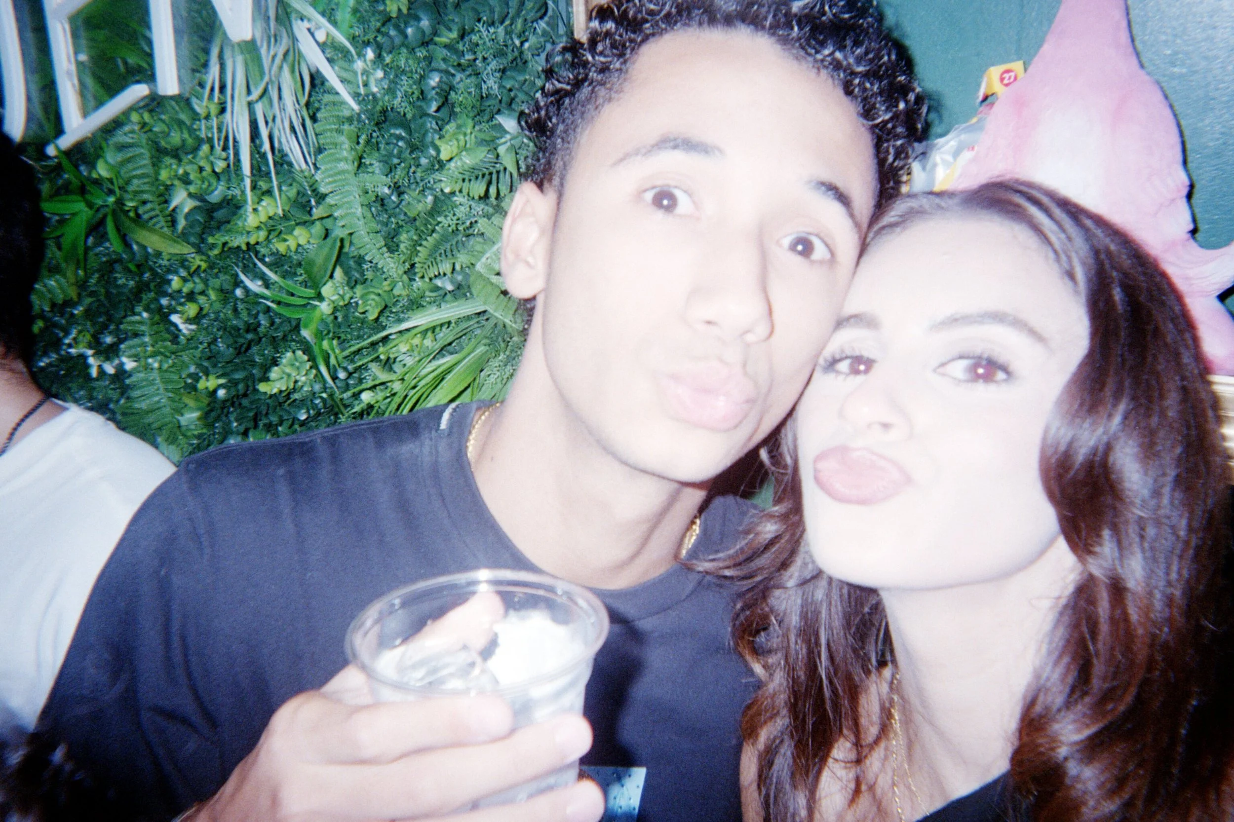 Two young adults, a man and a woman, are taking a close-up selfie together, making kissy faces. The man is holding a clear plastic cup, and there is greenery in the background.