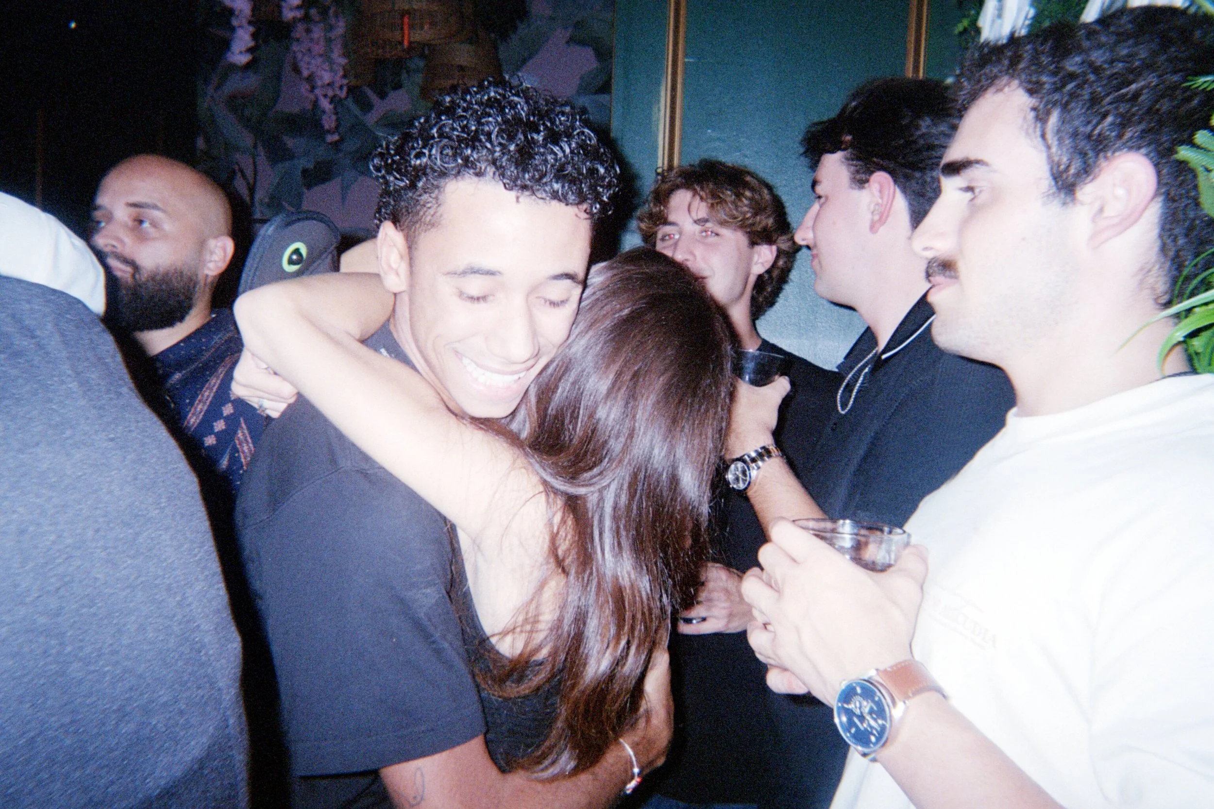 People hugging and socializing at a party or social gathering, with a man smiling and a woman embracing him.