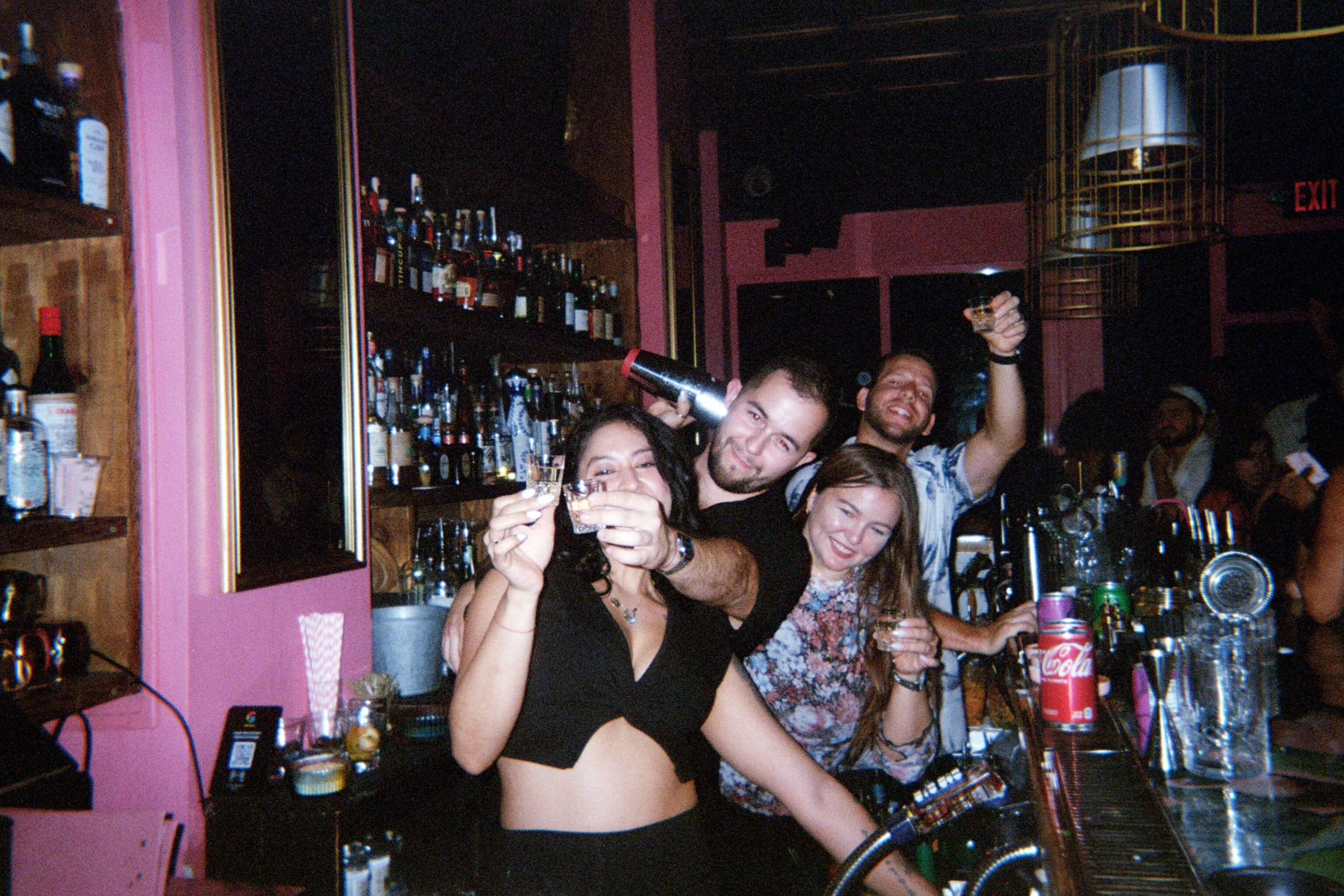 Four friends at a bar, smiling and holding drinks, with bartender behind the counter surrounded by bottles of alcohol.