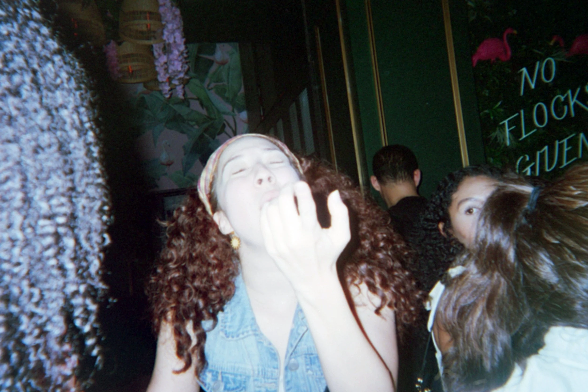 A woman with red curly hair, closed eyes, and a headband, covering her mouth with her hand in a crowded indoor setting with people and a sign that reads 'No Lock Garden.'