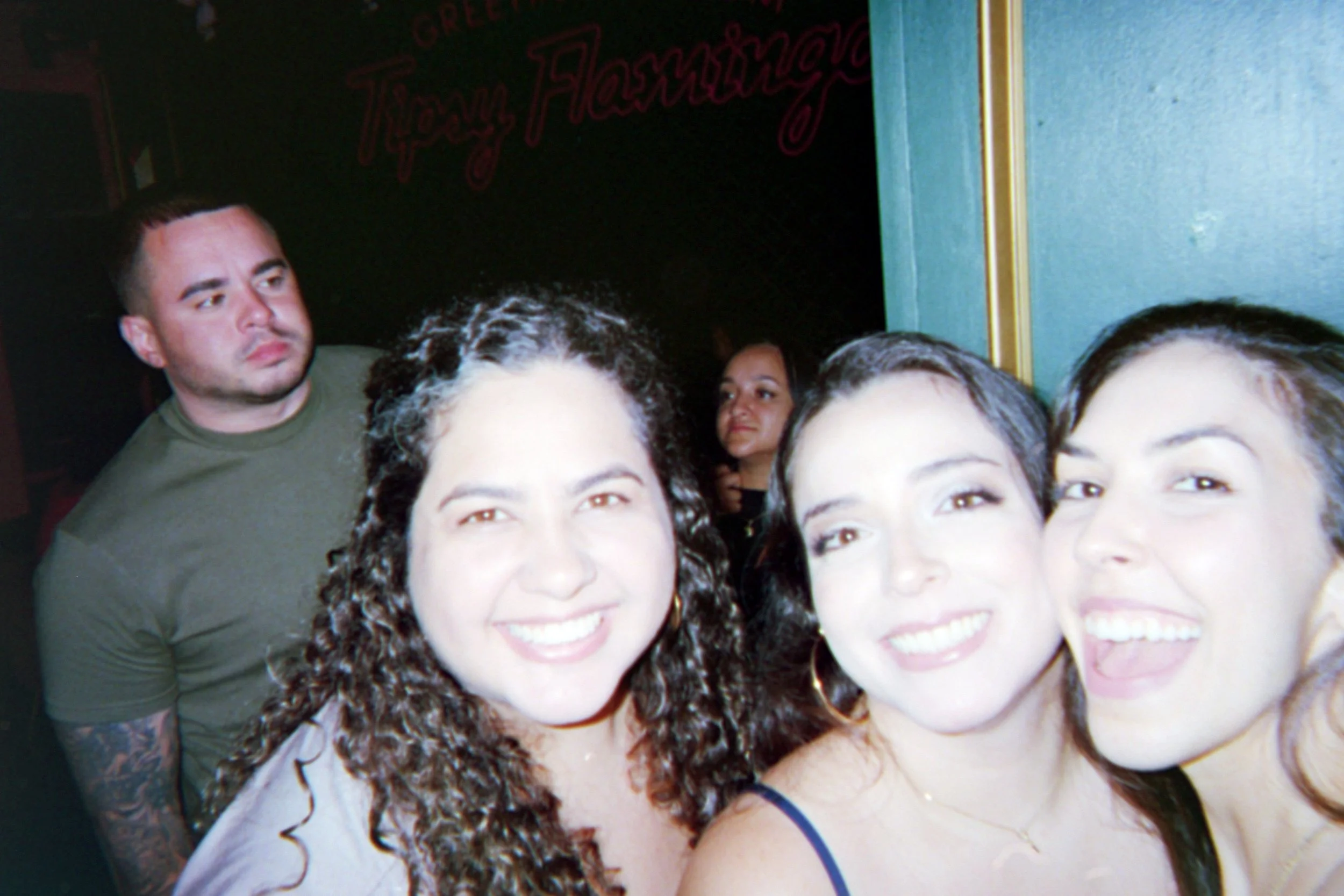 A group of four women smiling closely for a selfie, with a man in a green shirt standing in the background in a dimly lit environment, possibly a nightclub or pub.