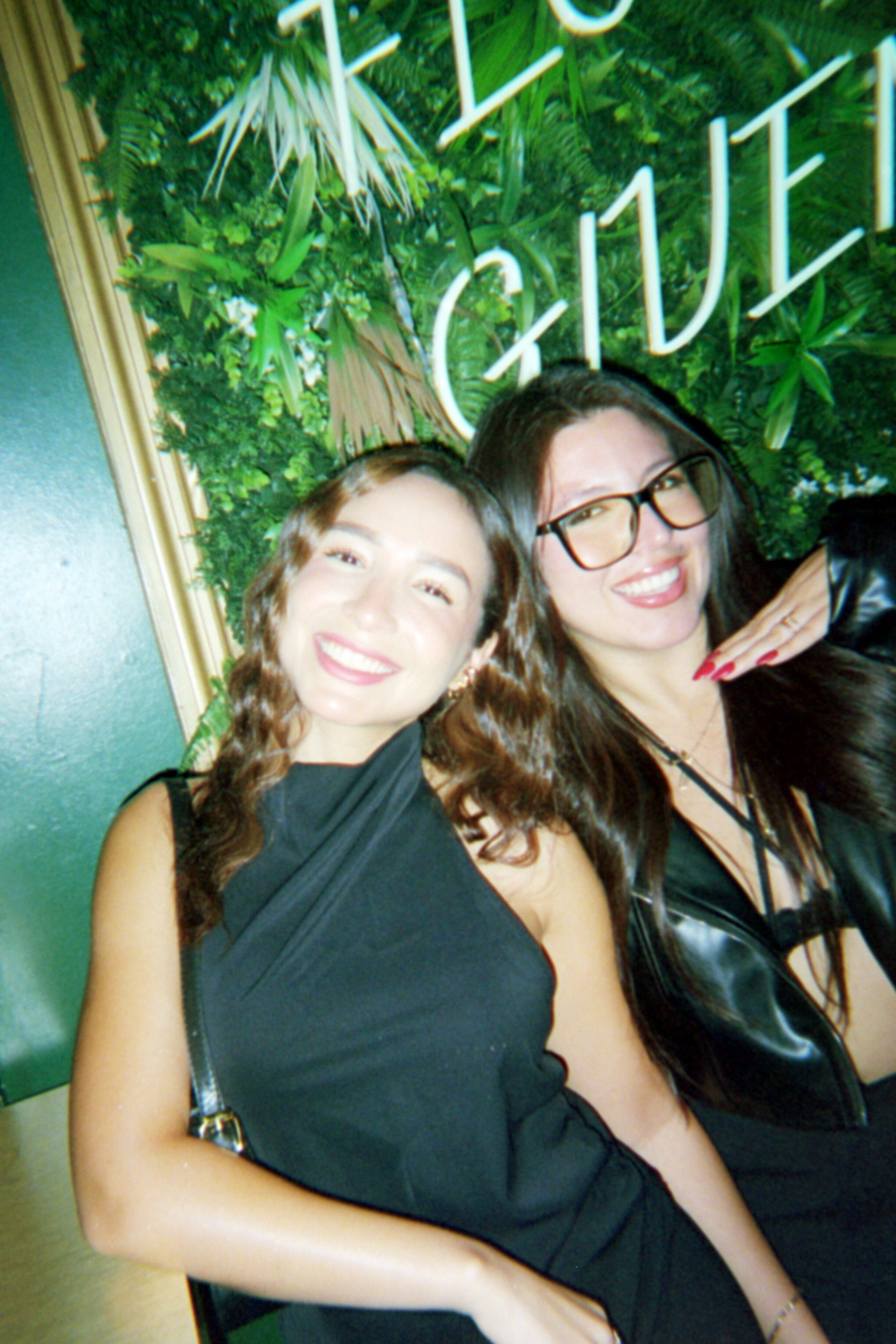 Two smiling women posing for a photo in front of a leafy green wall with illuminated white lettering that says 'GIVE'.
