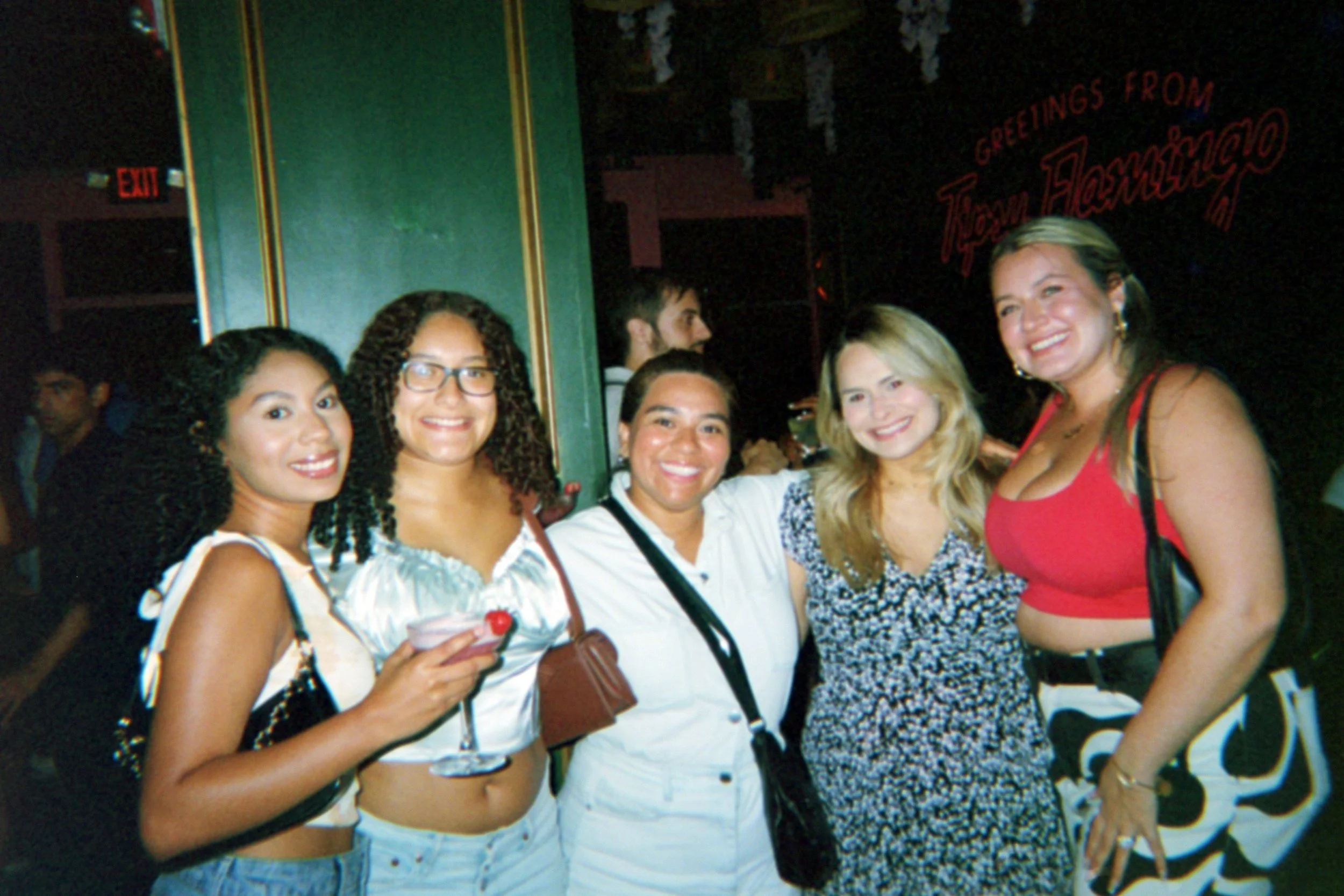 Five women smiling and posing together at a social event or bar, with a sign in the background that says "Greetings from Tampa, FL."