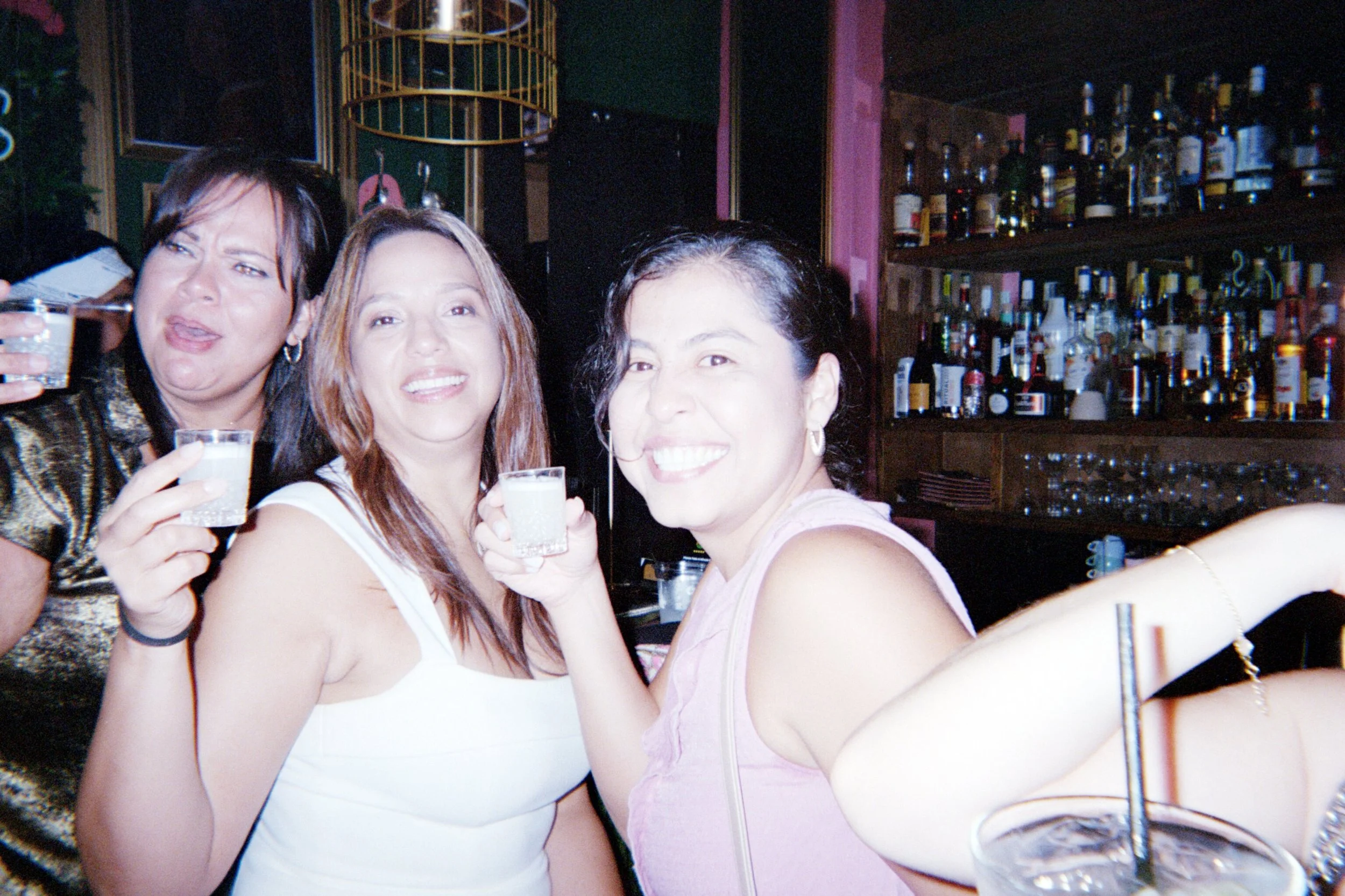 Three women smiling and holding shot glasses at a bar.