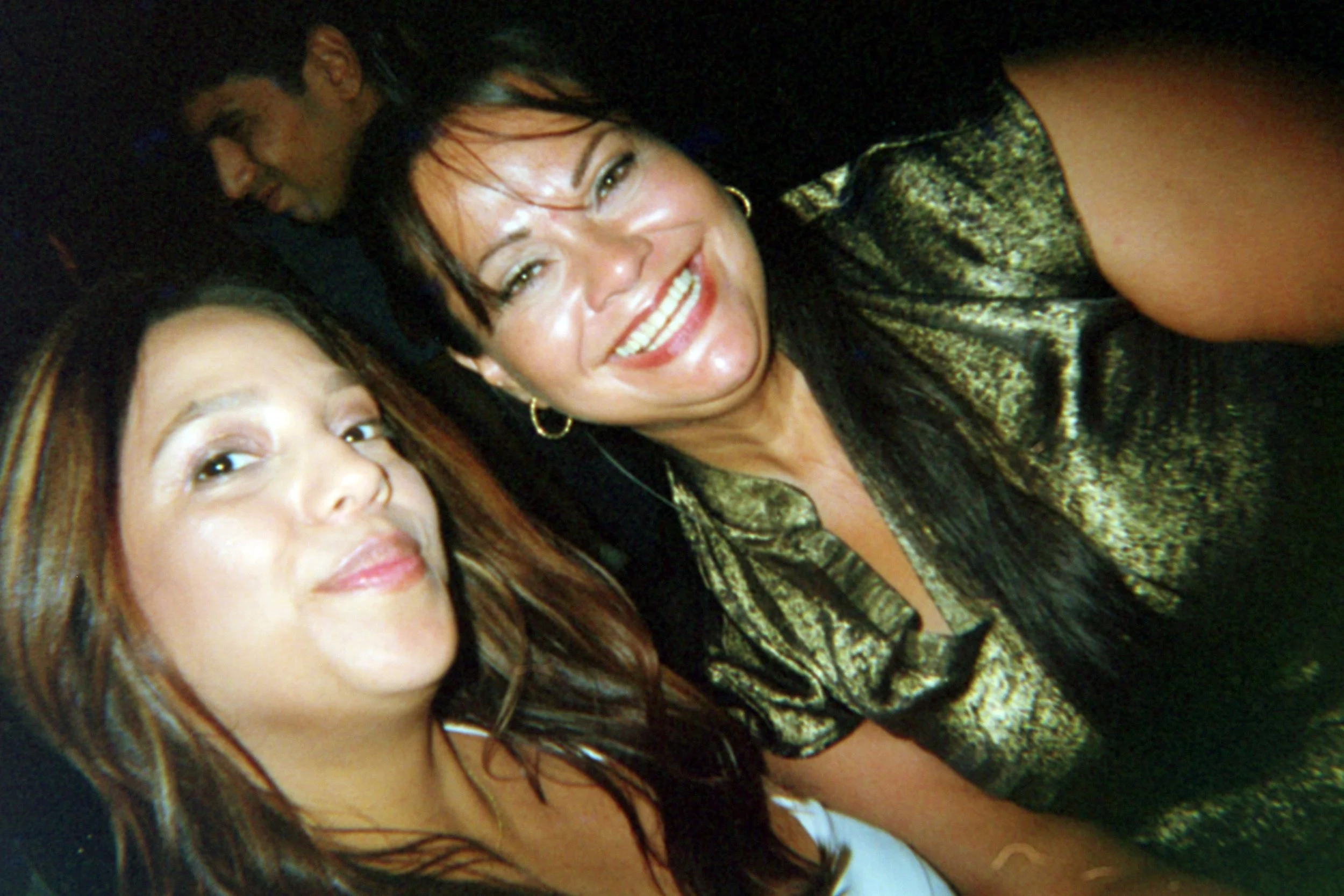 Two women taking a selfie, smiling, with a man in the background at a social event.