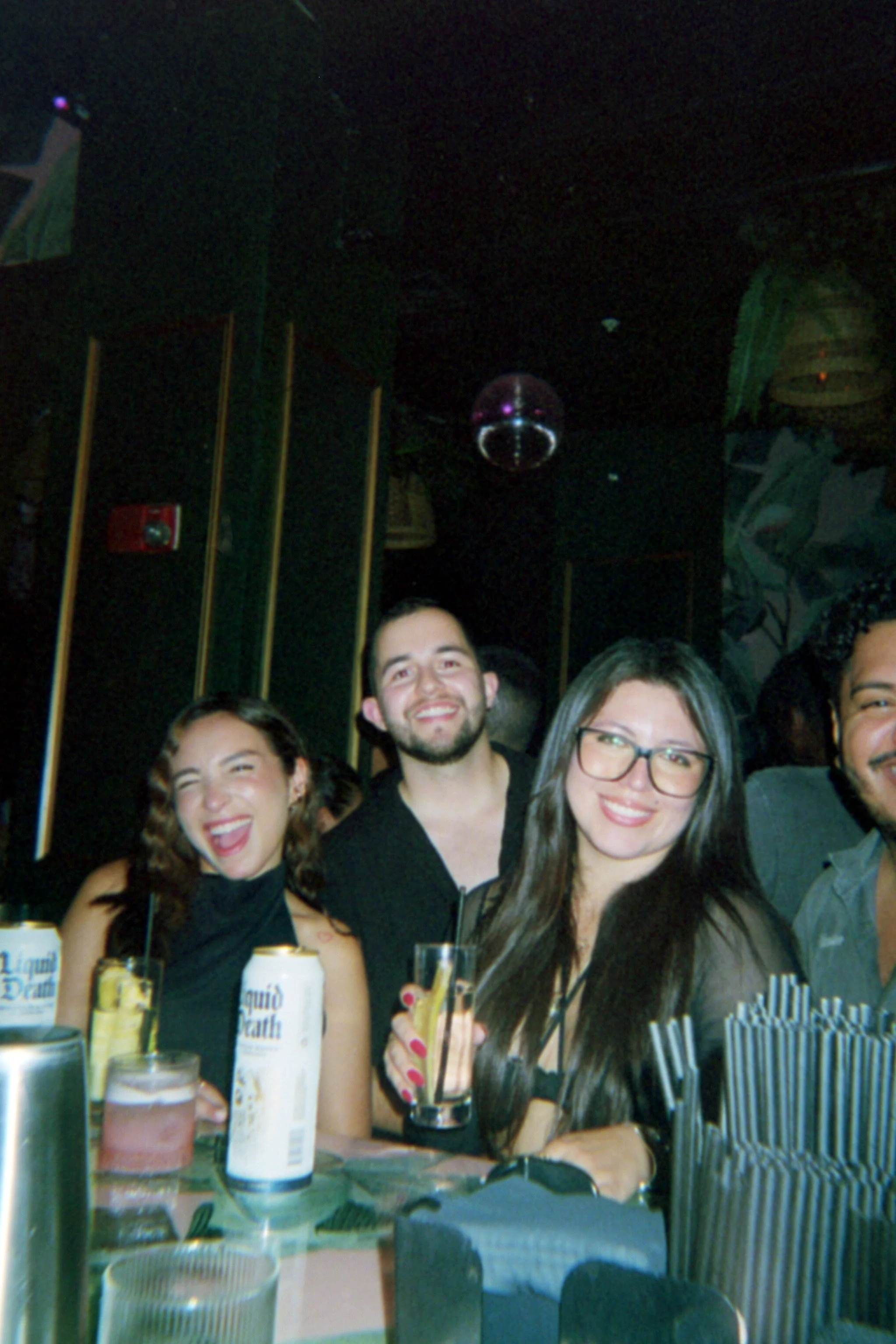 Group of friends enjoying drinks at a bar or club, smiling and laughing, with cans and glasses on the table.