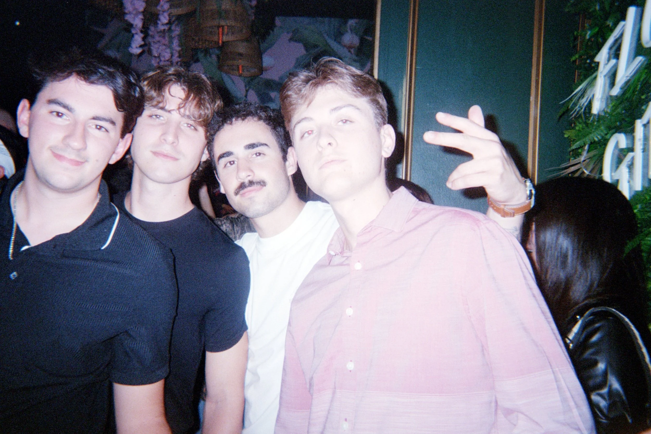 Four young men posing together at a social gathering or party, with some people visible in the background and a wall with greenery and decorative elements.