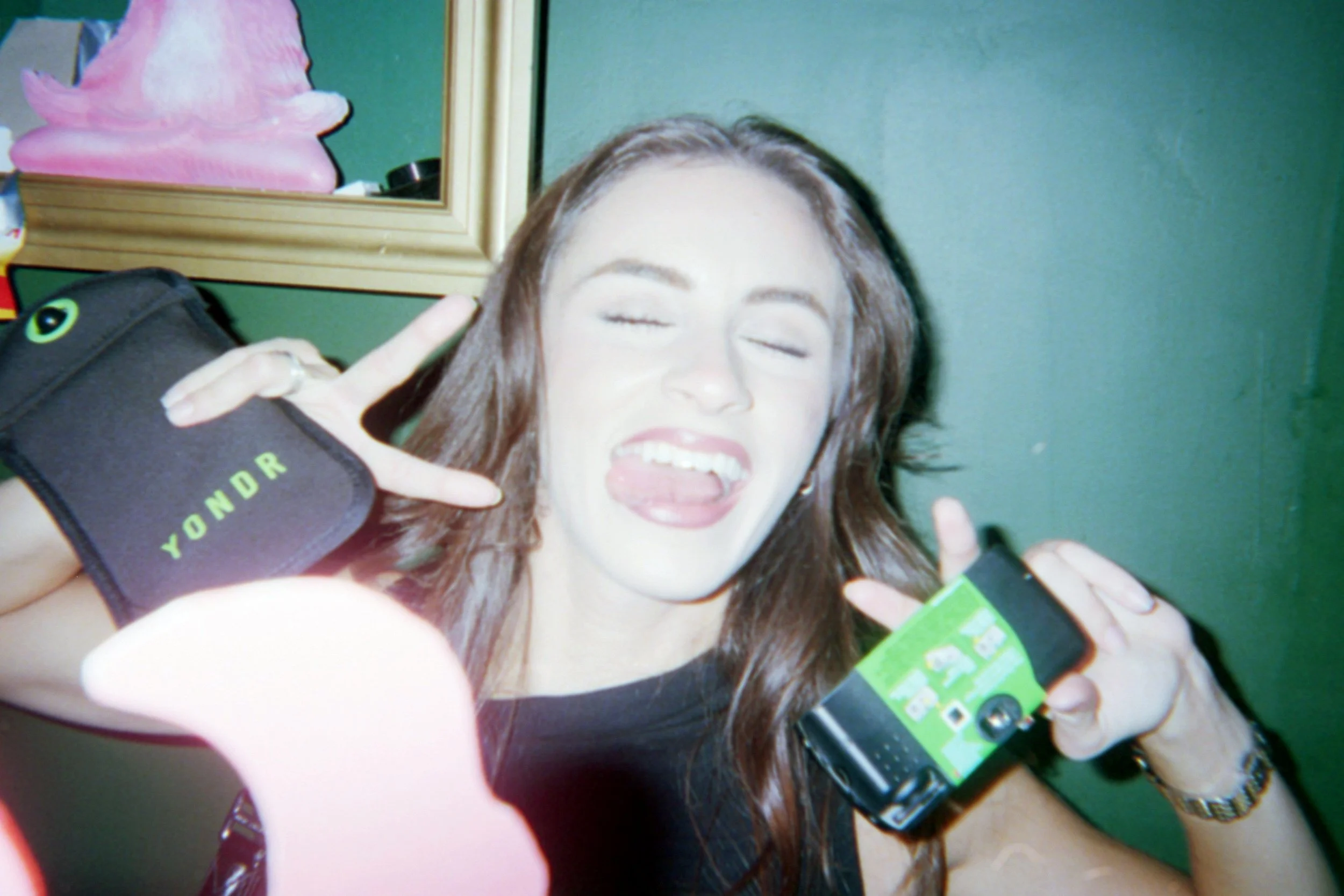 Woman with brown hair making a playful expression, holding a flip phone in one hand and a YOND R pouch in the other, with a green wall and mirror in the background.