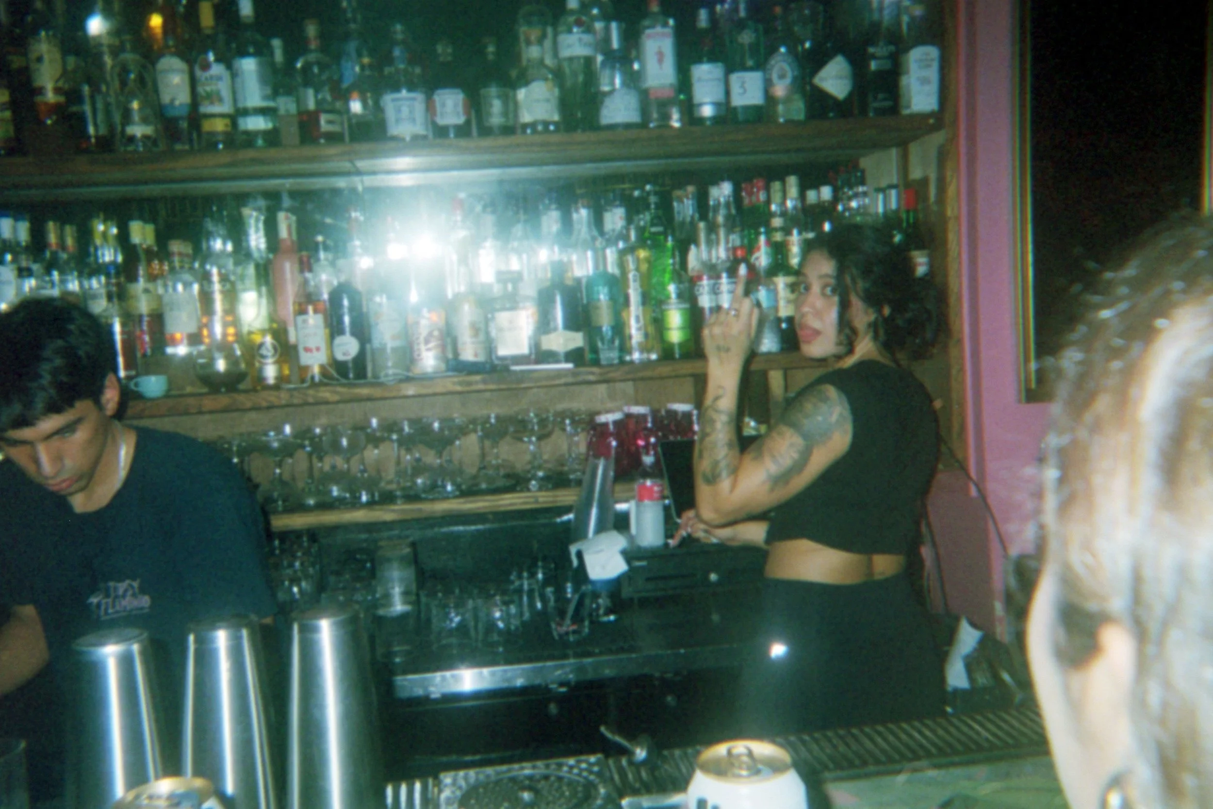 A woman with tattoos, dark hair, and a black top at a bar, looking over her shoulder. A bartender is visible on the left, and the bar shelf behind them is stocked with various bottles of alcohol.